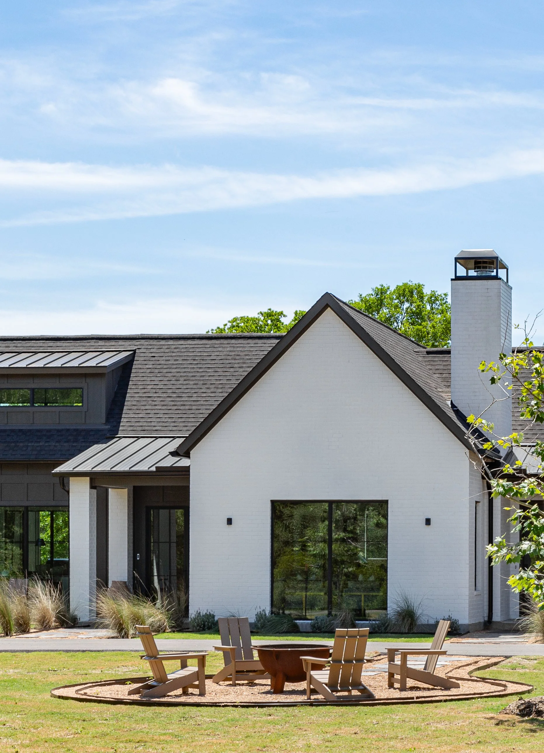 Exterior view of a modern house with white brick walls, black window frames, and a gray roof, surrounded by a lawn with a fire pit and four Adirondack chairs.