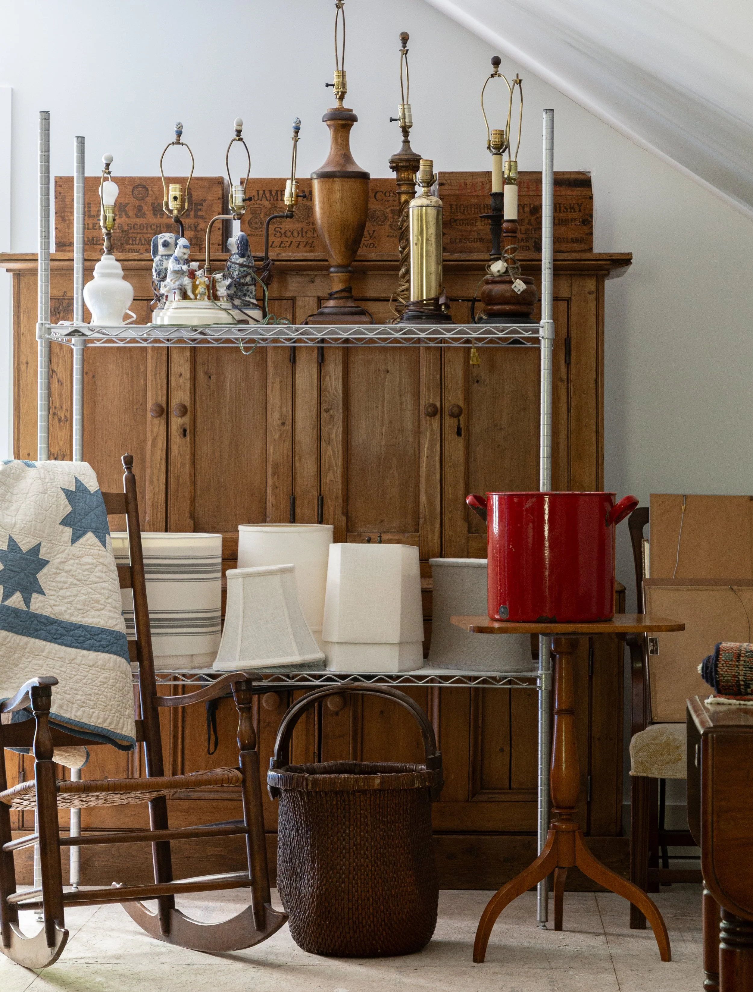 An indoor scene with a wooden cabinet, a metal shelving unit with vintage lamps and ceramic figurines, a basket, a red pot on a small wood table, and various lampshades and household items.