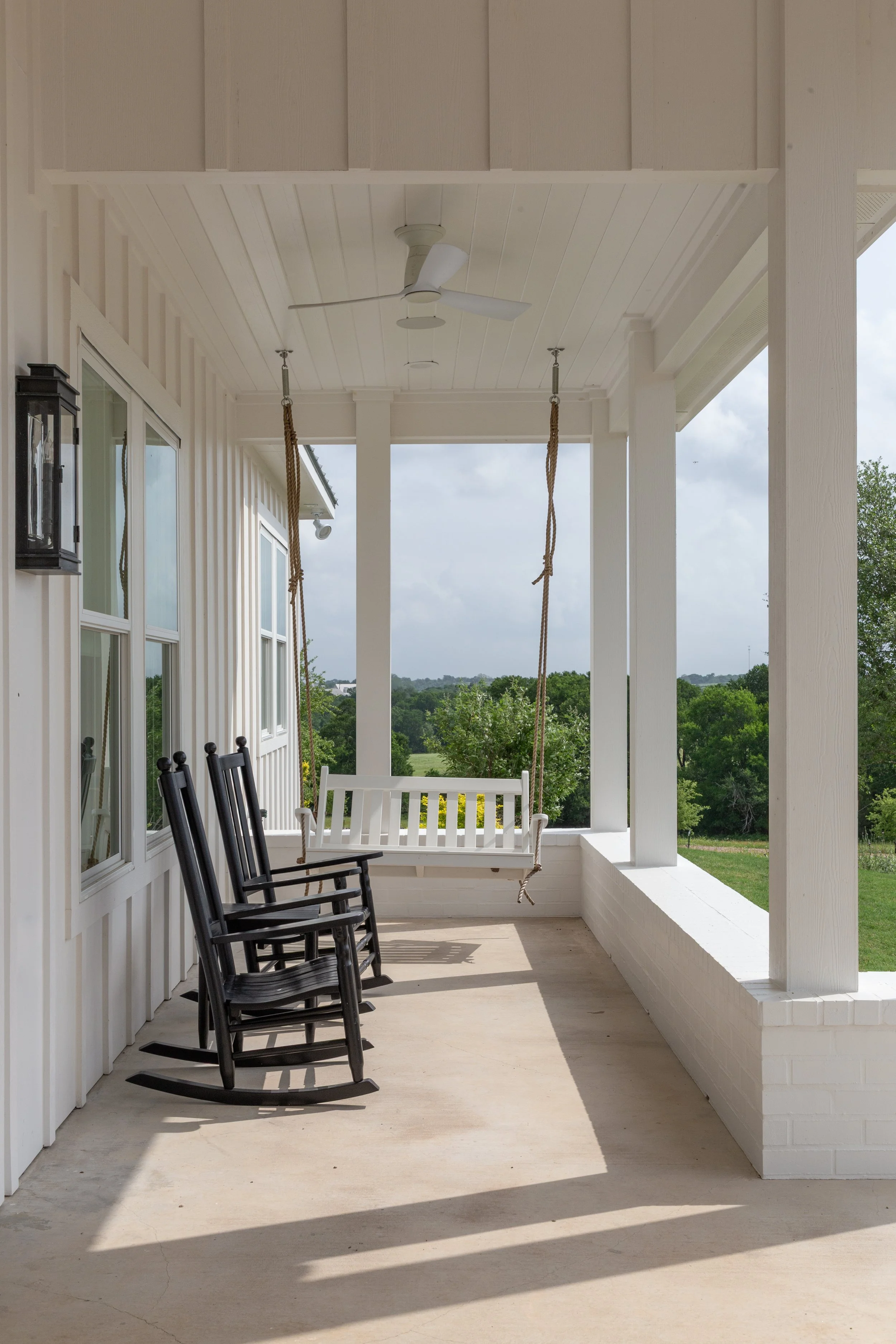 A covered porch with white walls and ceiling, featuring a ceiling fan, black and white rocking chairs, and a white porch swing hanging from the ceiling. Green landscape visible in the background.