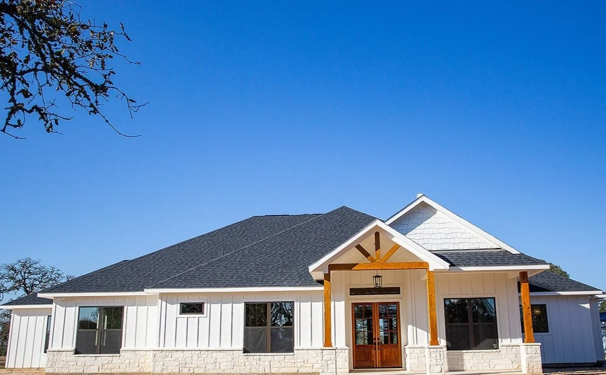 Front view of a white house with a black roof, wood accents, and a stone foundation, under a clear blue sky.