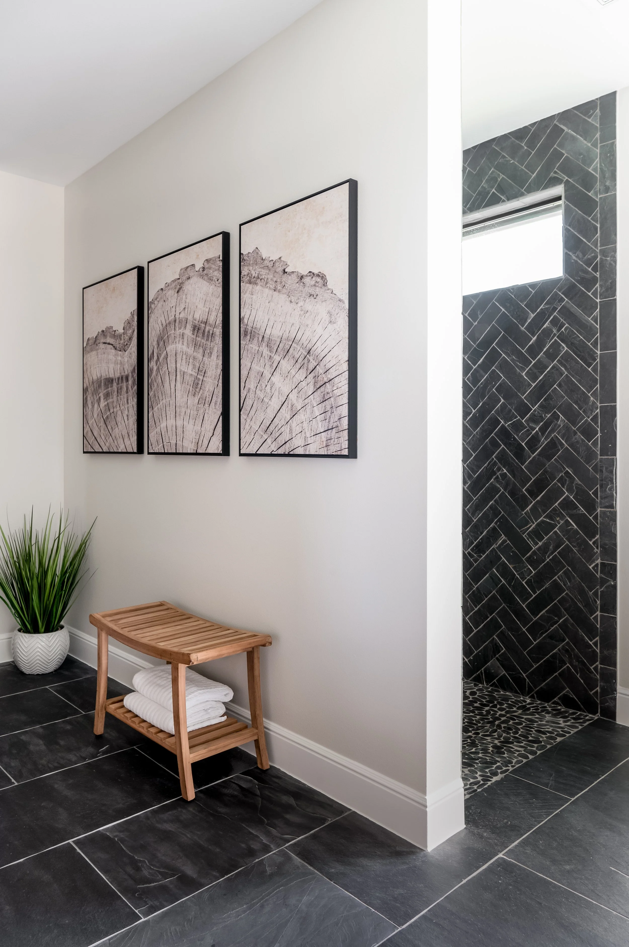 Interior bathroom corner with black slate floor, a white wall decorated with three framed art pieces, a small wooden bench with towels, a green plant in a white pot, and a walk-in shower with black tiling and a narrow window.