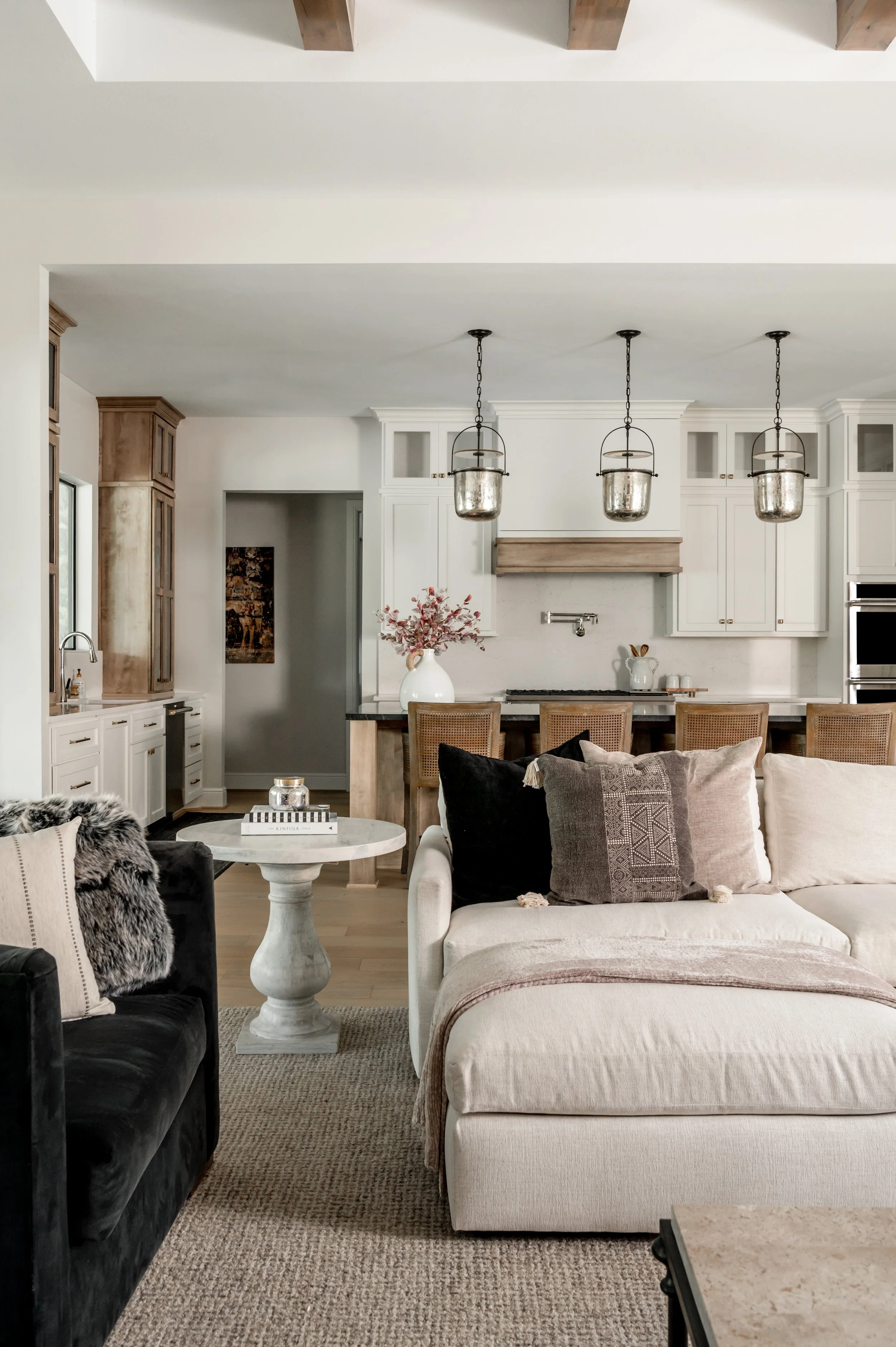 A living room with a white sofa, black accent chair with a faux fur throw, a round white side table with books and candles, and rustic pendant lighting over the kitchen area in the background, featuring white cabinets and wooden accents.