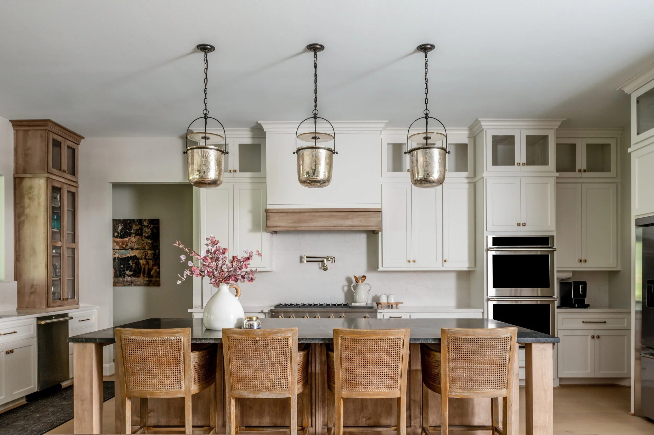 Modern kitchen with white cabinets, a wooden island with six cane-back chairs, three metallic pendant lights, a vase with pink leaves, and stainless steel appliances.
