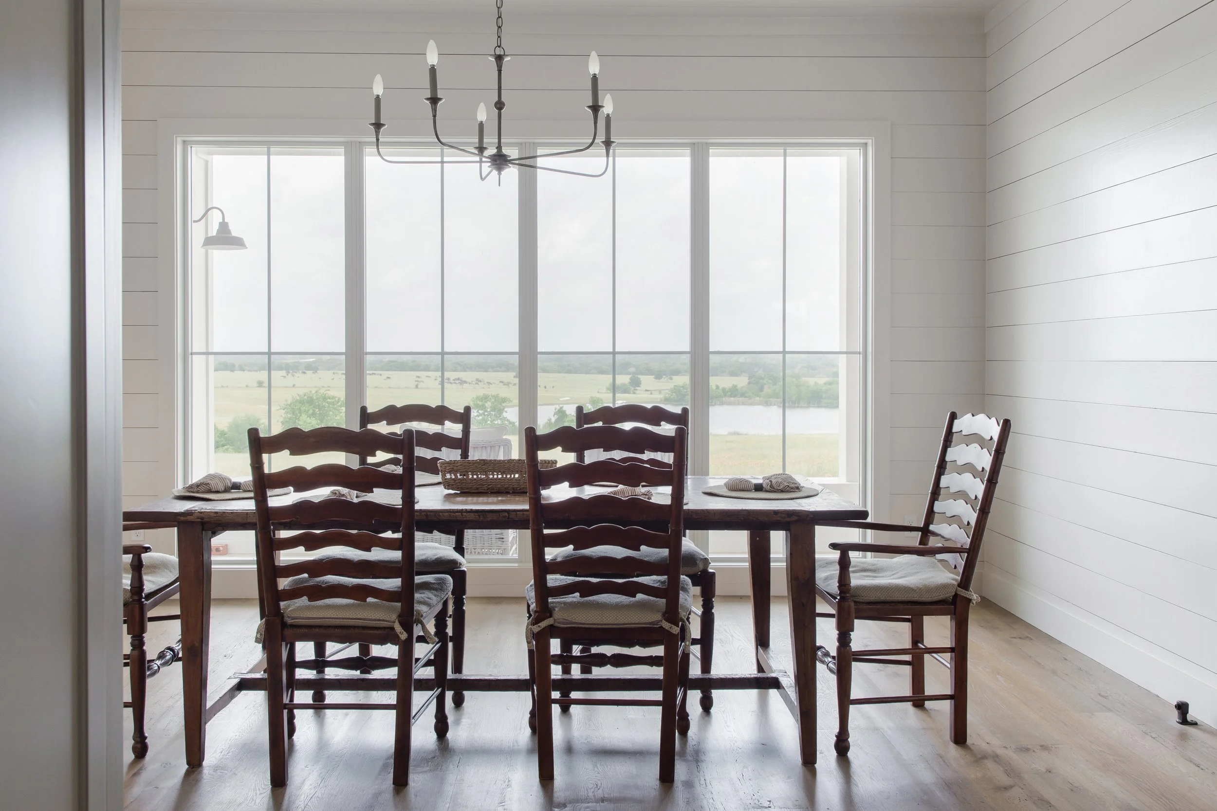 Dining room with a large window view of a rural landscape, wooden dining table, six wooden chairs, two place settings, and a rustic chandelier hanging from the ceiling.