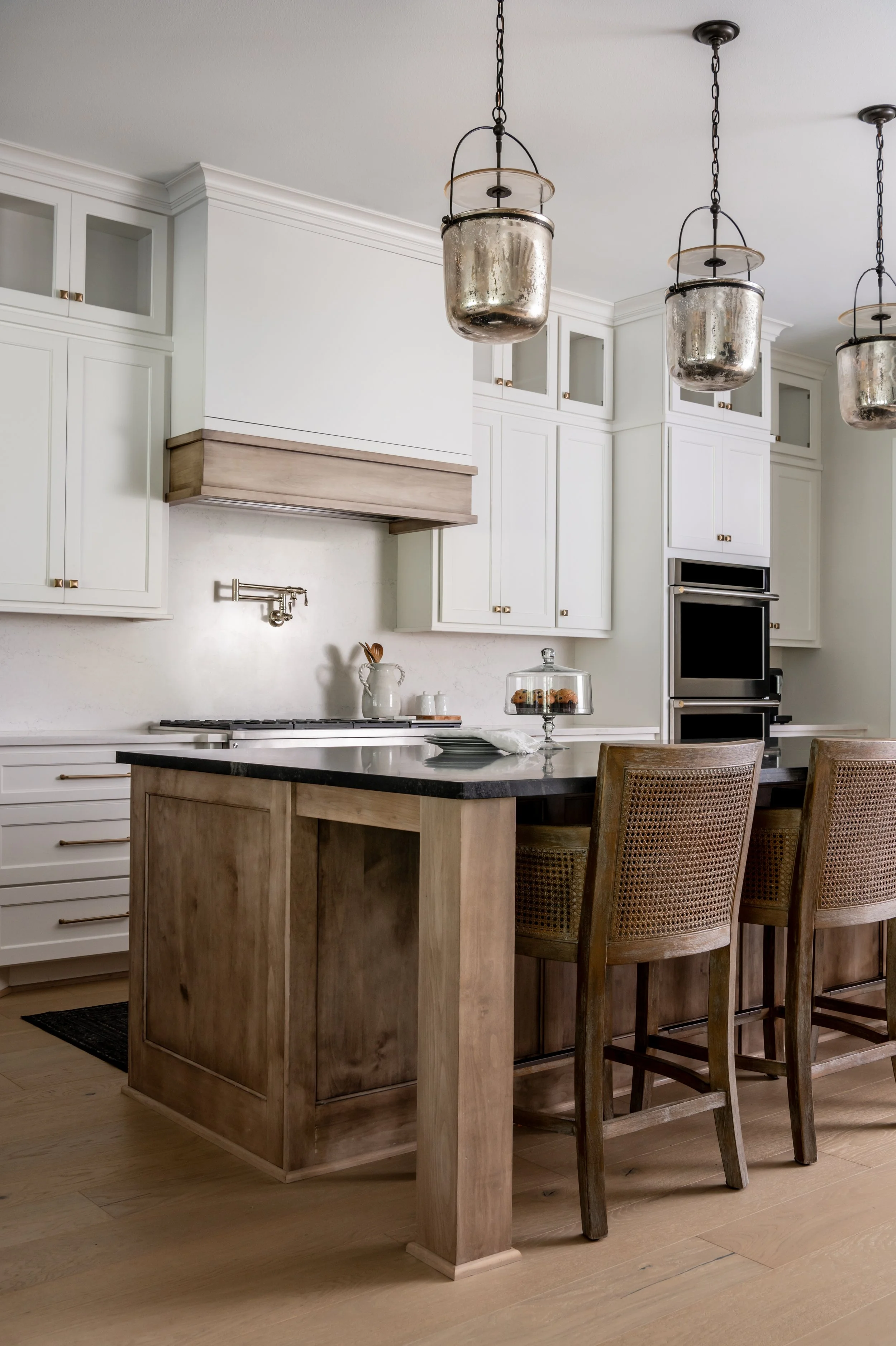 Contemporary kitchen with white cabinets, a central island with a black countertop, wooden chairs with woven backs, and three metallic pendant lights hanging from the ceiling.