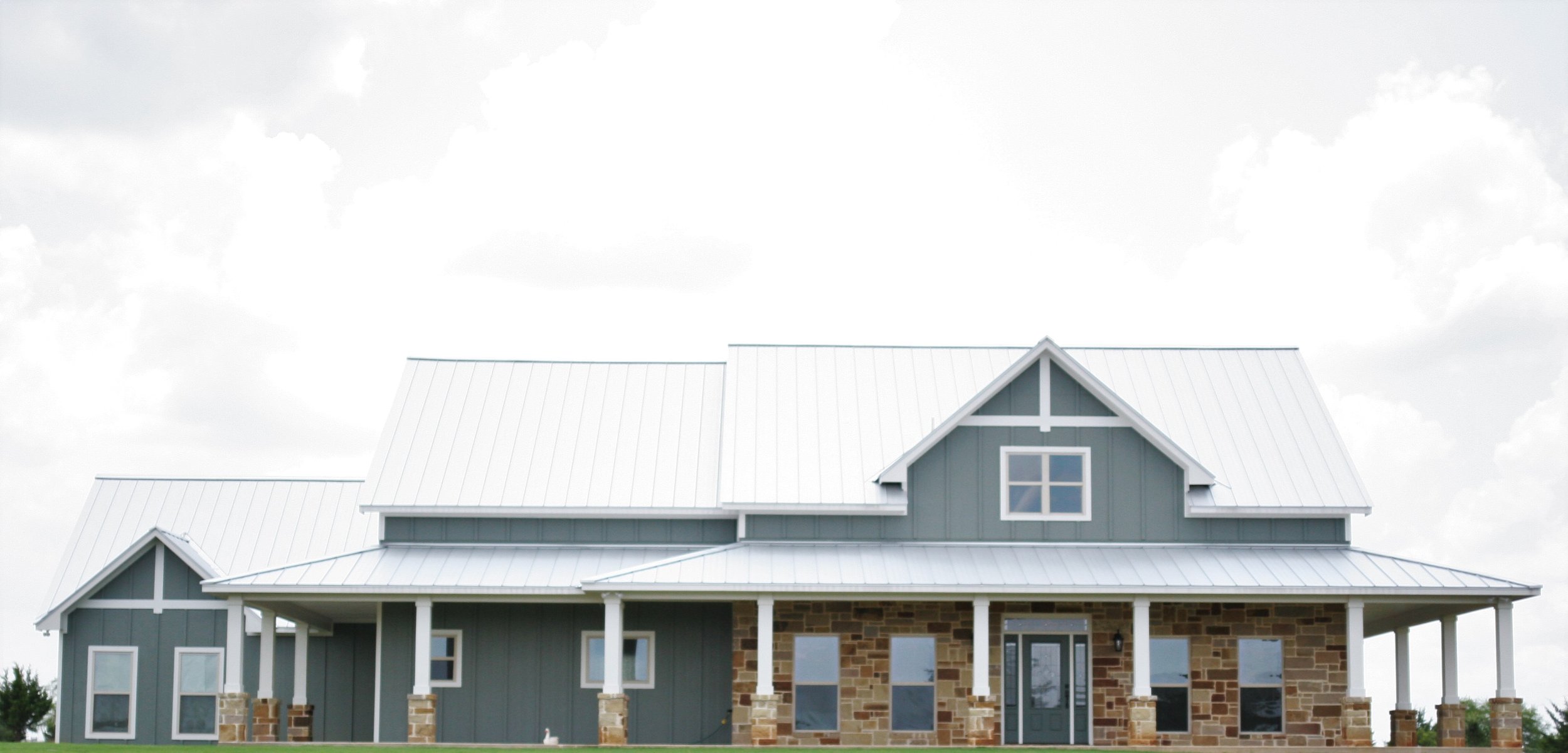 A two-story house with a metal roof, gray siding, stone accents, and multiple windows, including a dormer window, set against a cloudy sky.