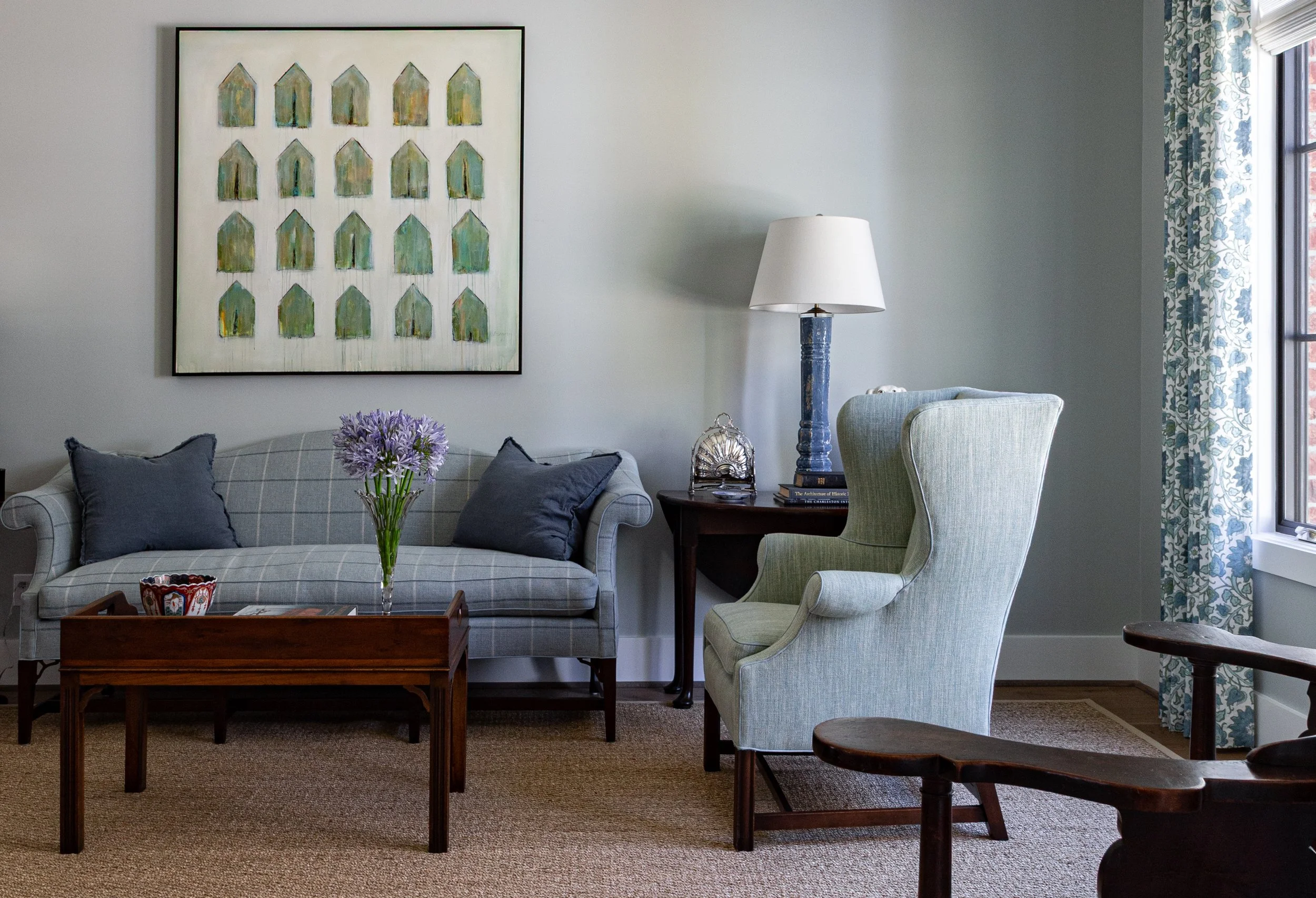 Living room with blueish-gray sofa, purple flowers in a vase, side table, lamp, armchair, and window with patterned curtains.