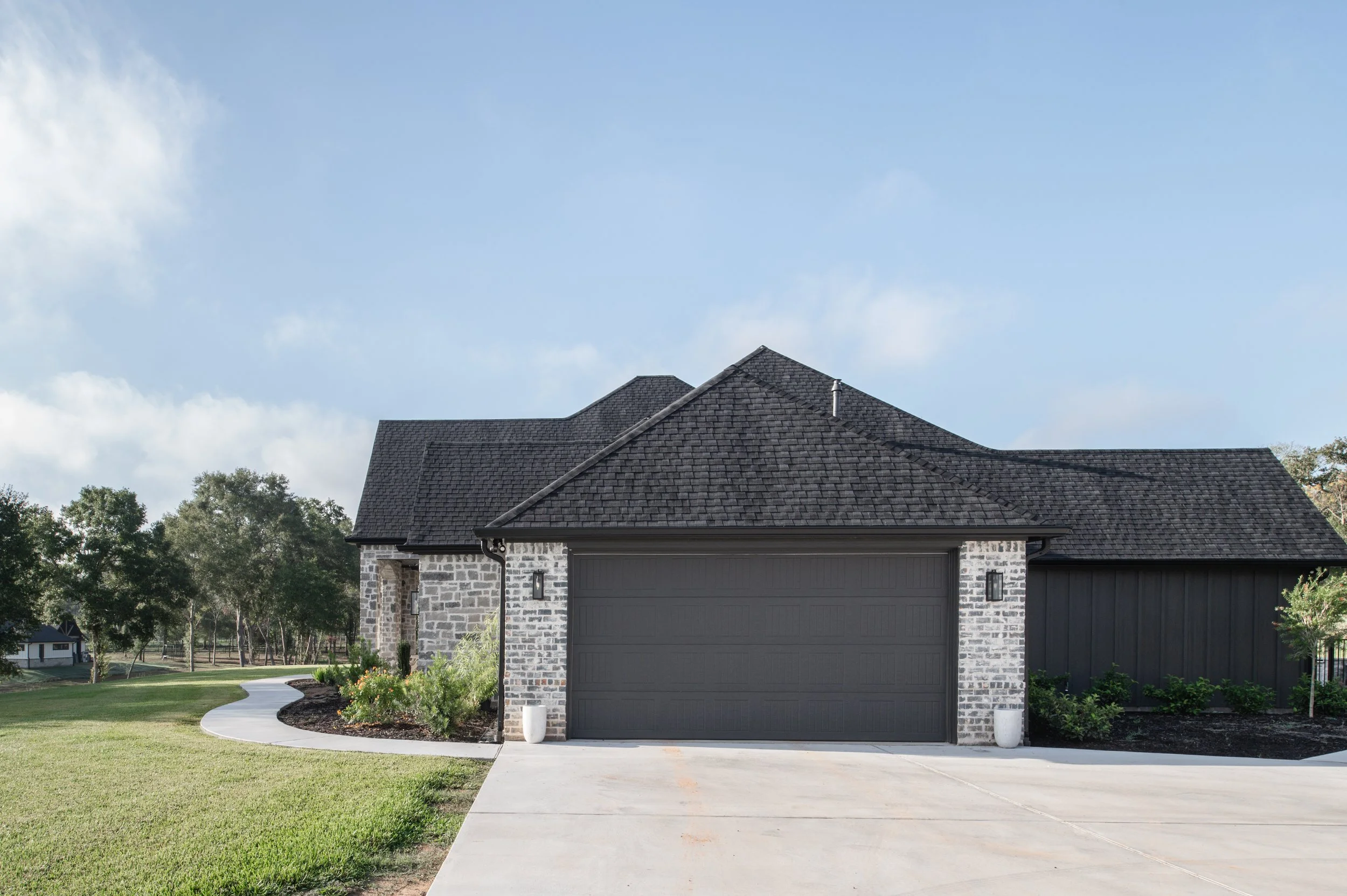 Modern house with black roof, gray brick facade, closed black garage door, and sidewalk leading to the entrance, surrounded by a green lawn and trees.
