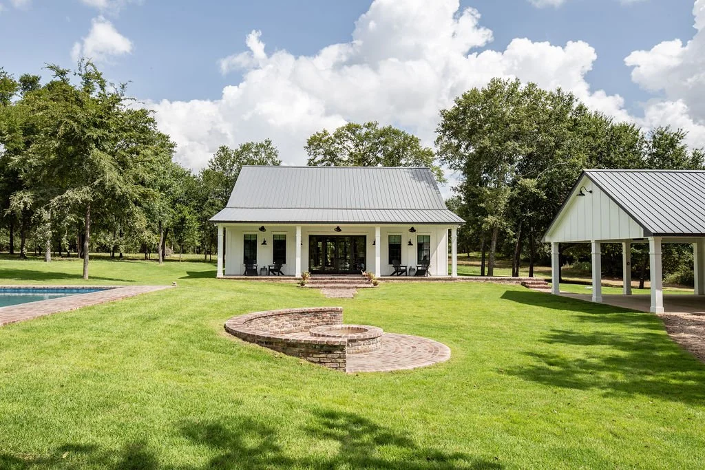 A white house with a gray metal roof surrounded by green lawn, trees, and a brick fire pit, with a small covered pavilion on the right and a swimming pool on the left.