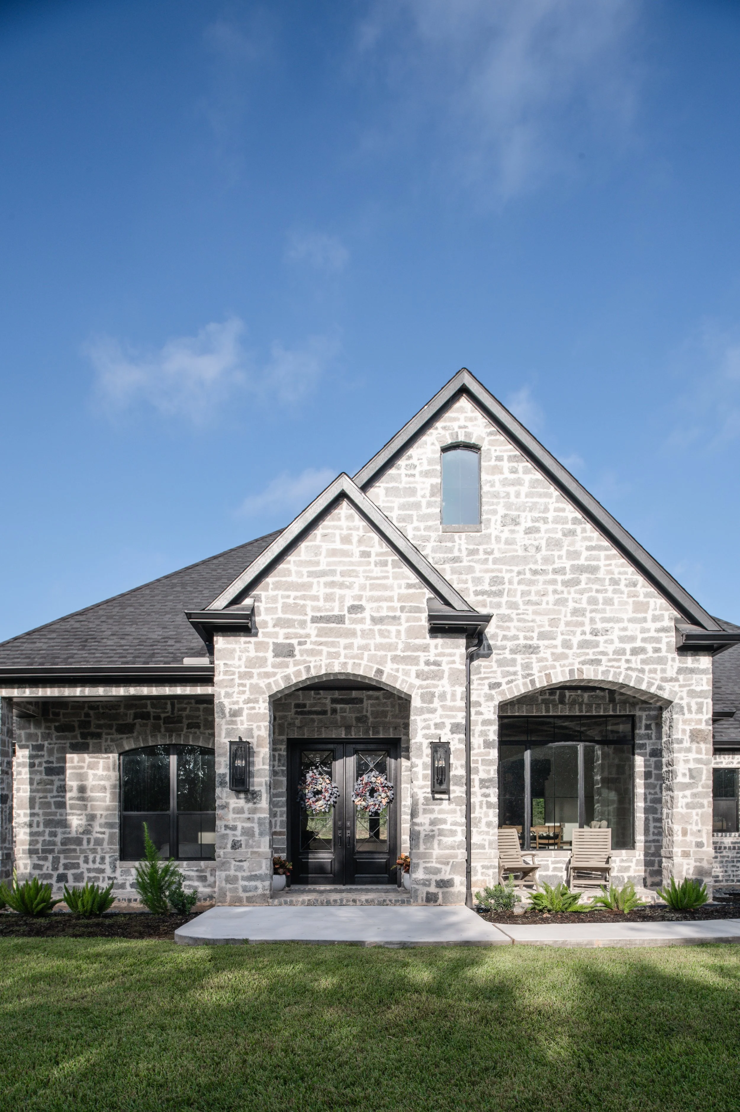 Front view of a modern stone house with a gray roof, black front door with wreaths, two windows, outdoor seating, and green lawn under a bright blue sky.