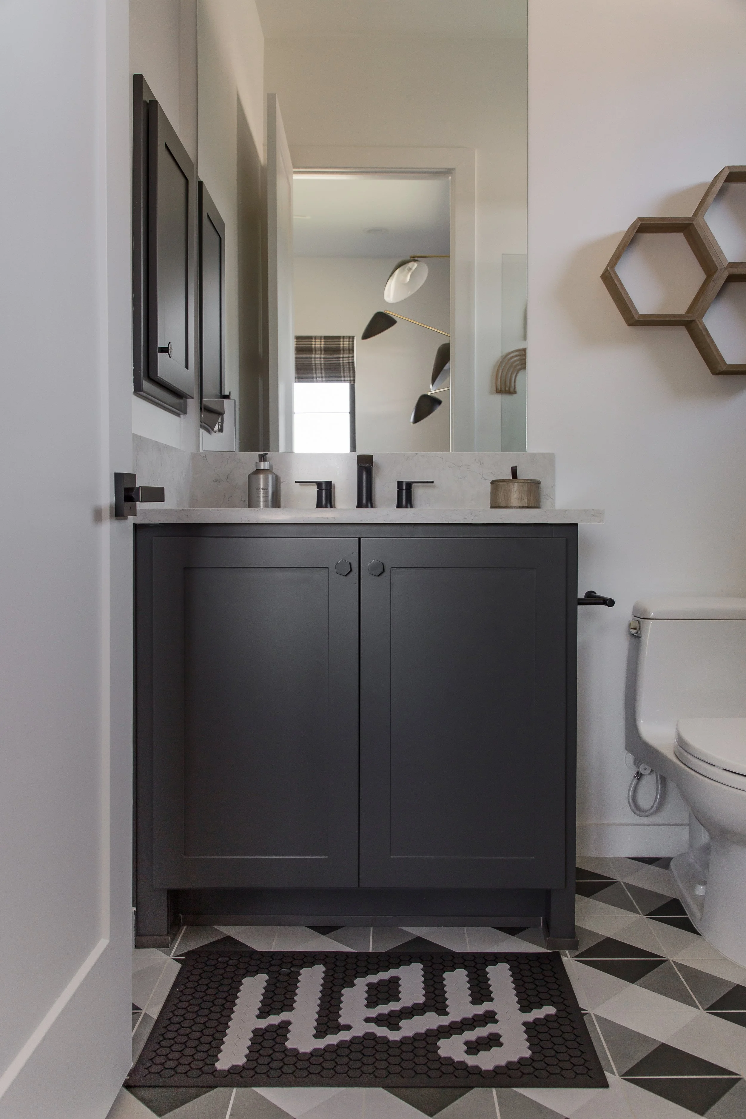 A modern bathroom with a black vanity, white marble countertop, and a large mirror. There is a towel holder with a towel and decorative hexagon shelves on the wall. A toilet is visible on the right, and a doormat with the words 'Hey' is on the patter