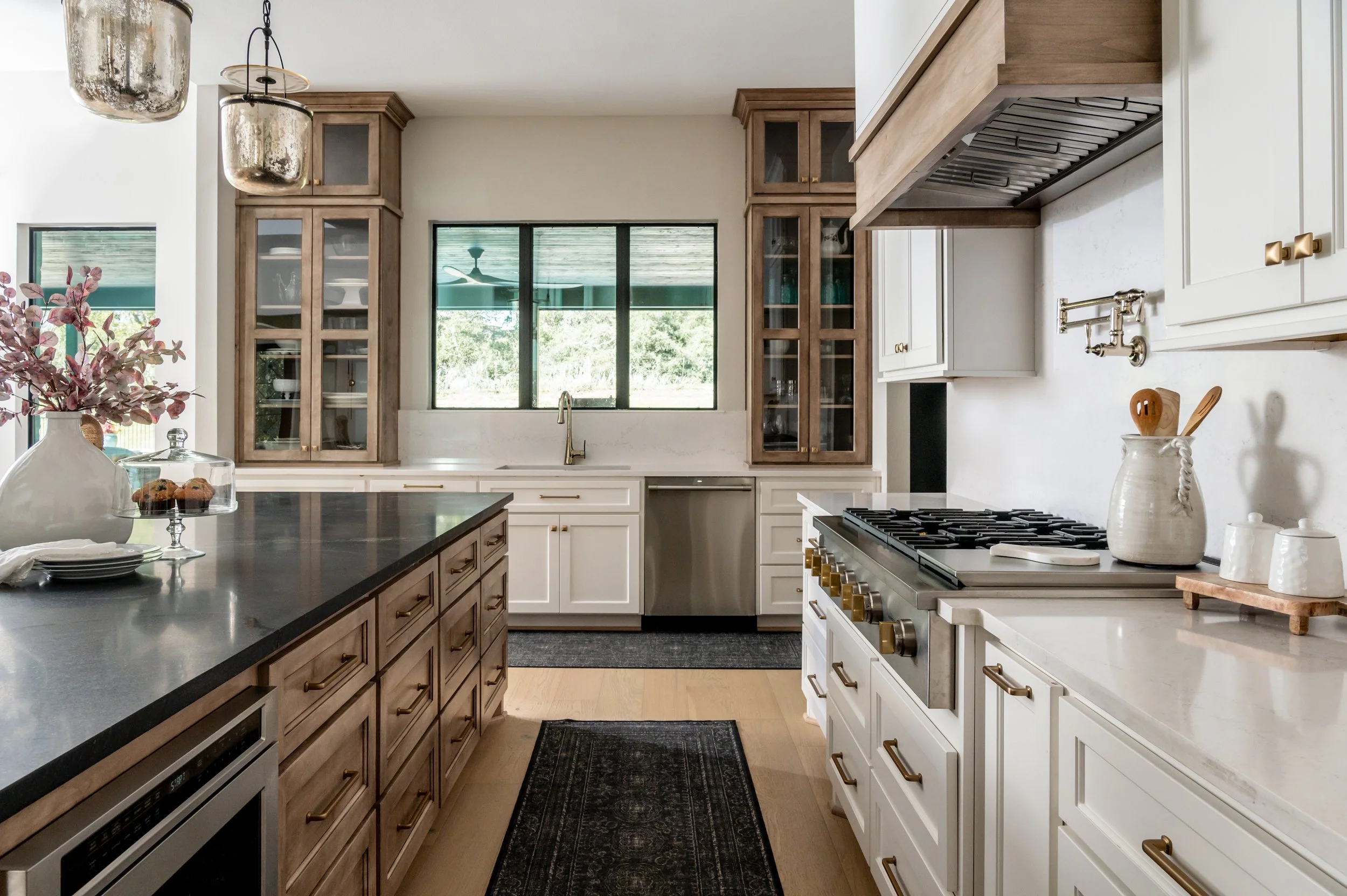 Modern kitchen with white and wood cabinetry, black countertops, stainless steel appliances, and a black rug on light wood flooring.