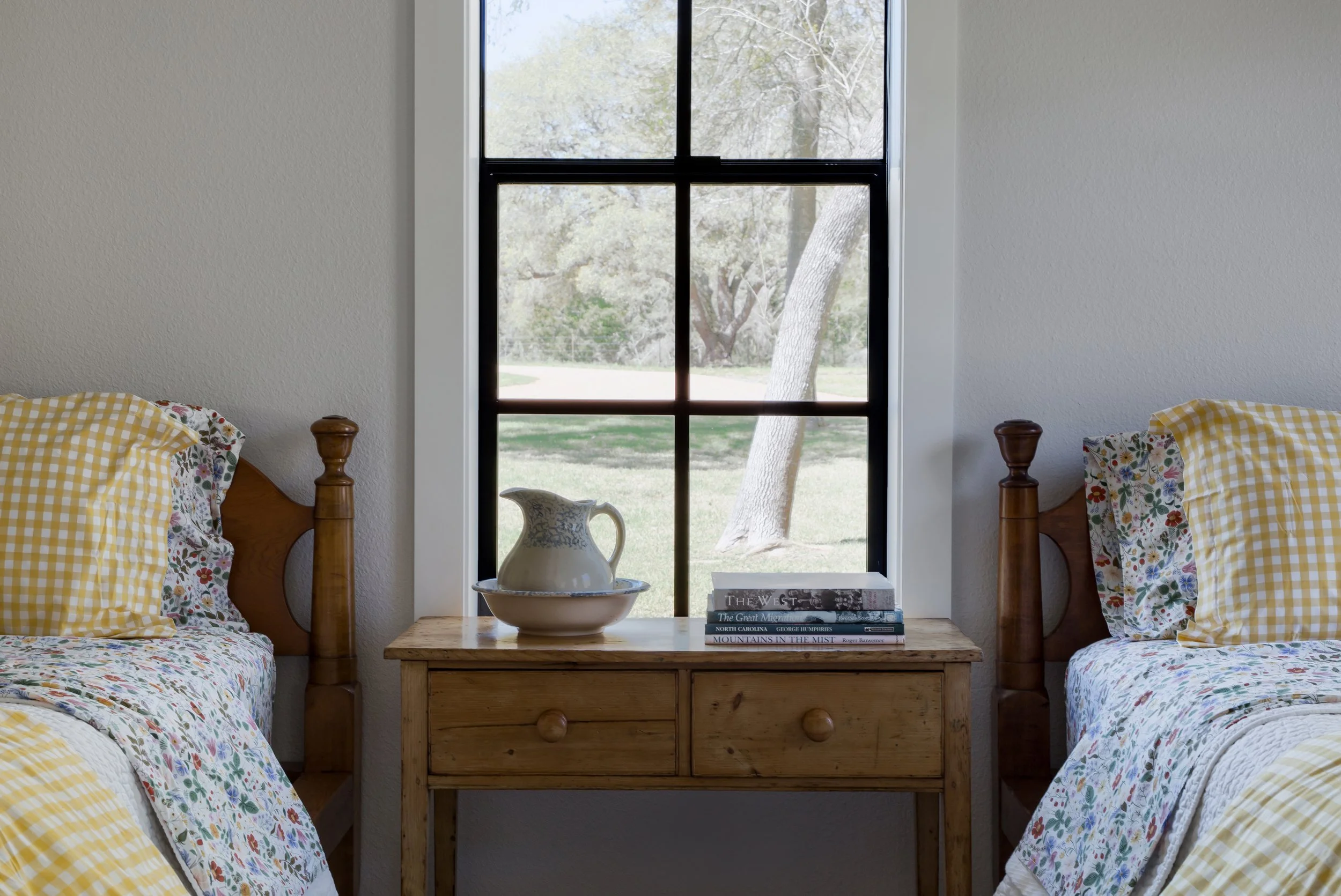Two beds with yellow and floral bedding separated by a wooden nightstand, in front of a window showing outdoor trees and grass.