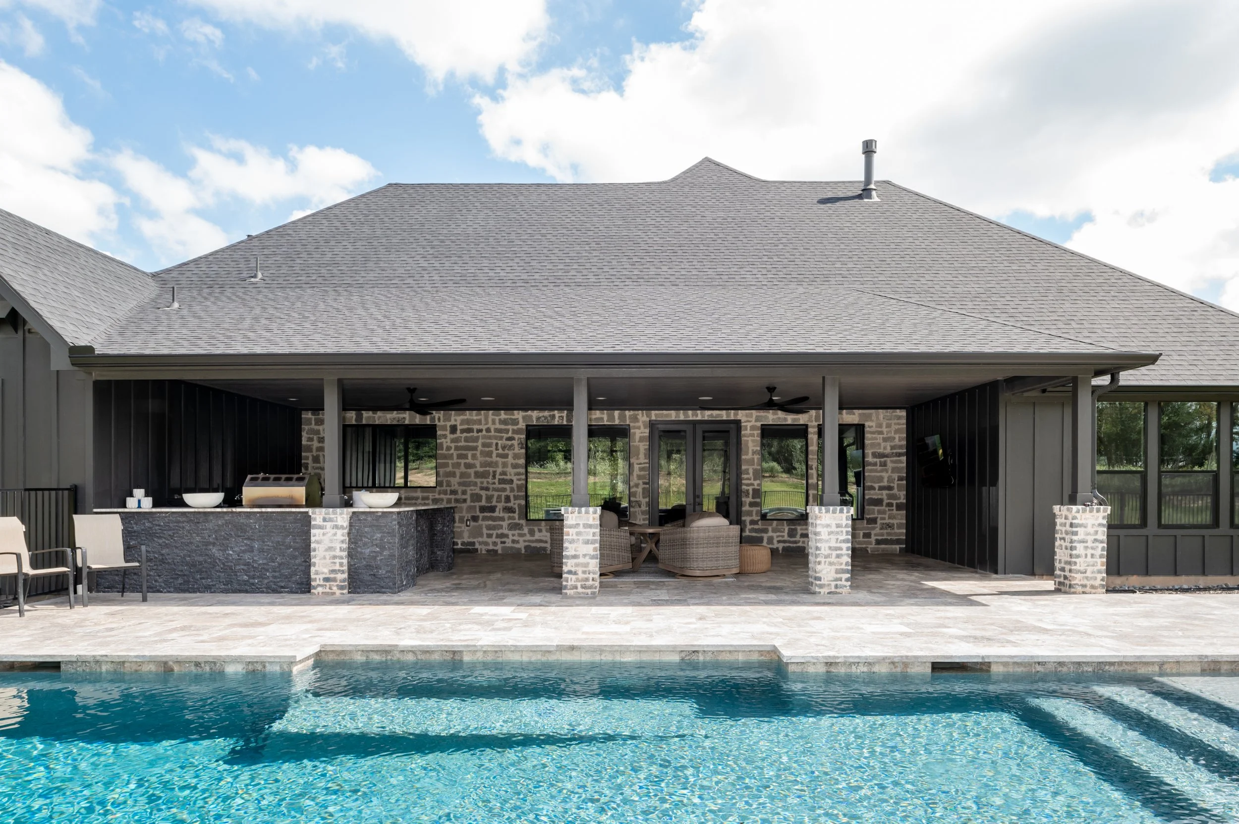 Backyard with a swimming pool and a covered outdoor lounge area with chairs, a table, and a built-in grill with double sinks, stone accents, and a brick wall, under a gray-shingled roof.