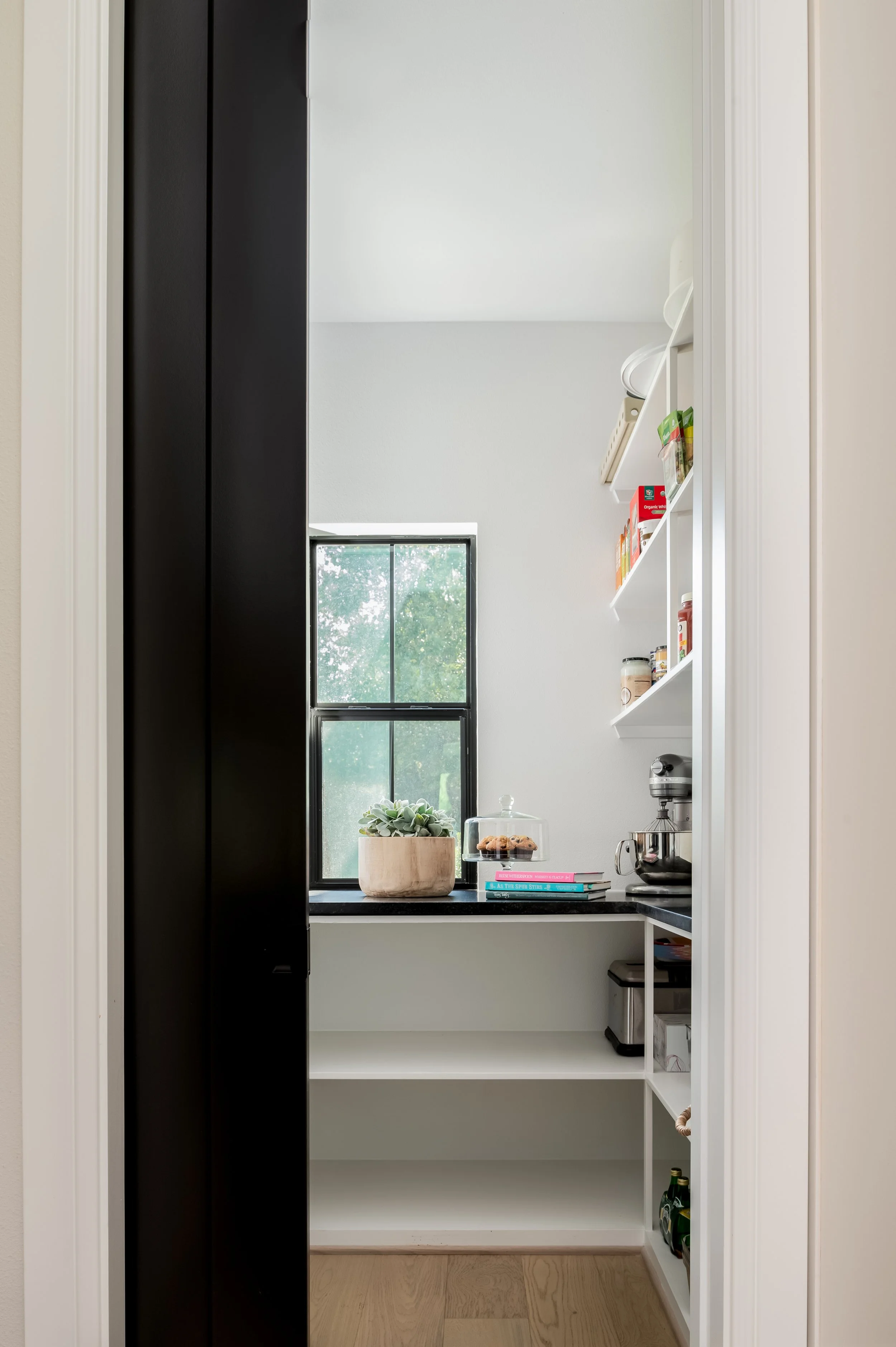 View into a small kitchen or pantry with white shelves, black countertop, and a window with greenery outside. On the counter, there is a plant in a wooden pot and a glass cake dome with cookies.