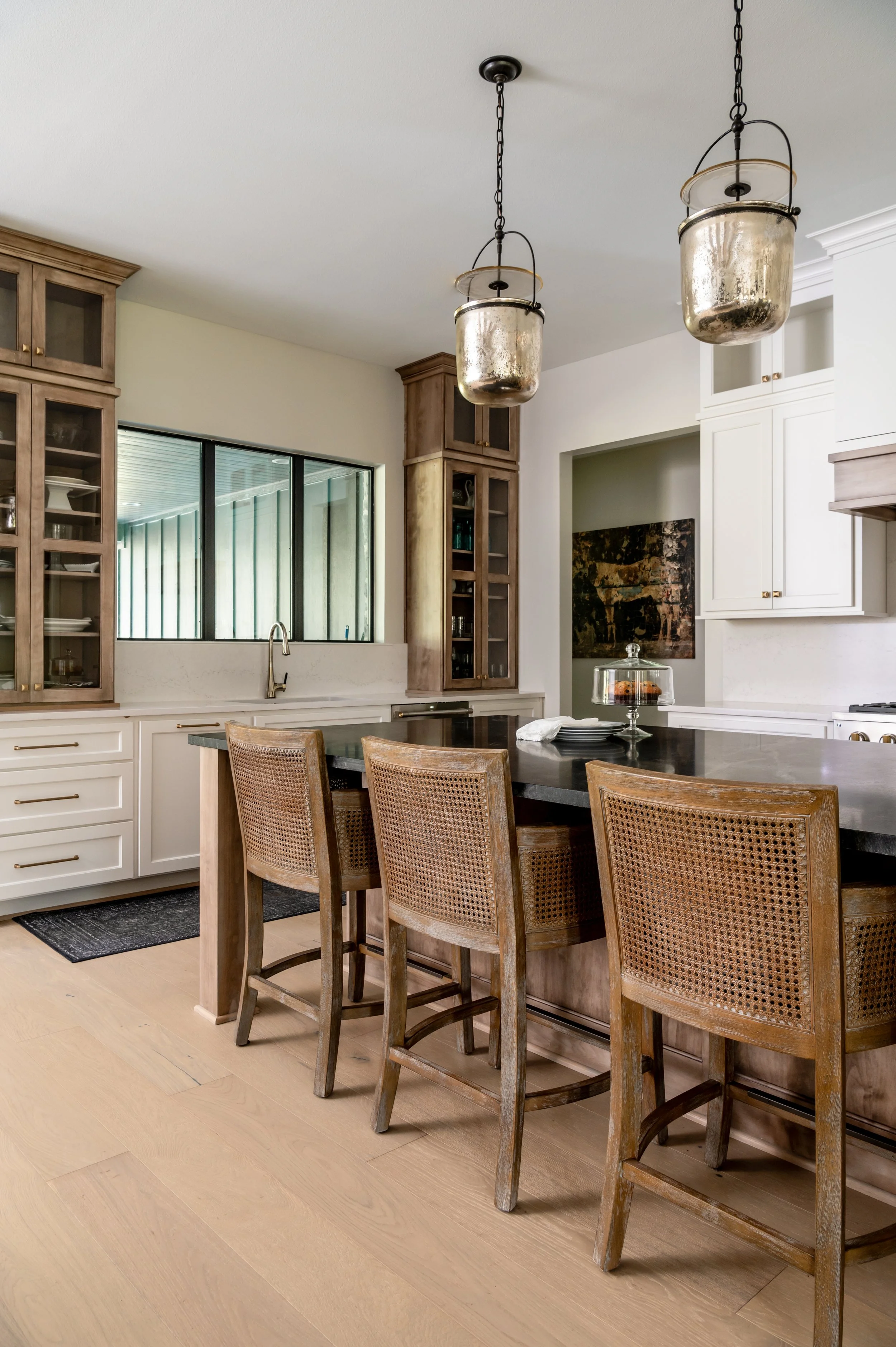 Kitchen with white and wood cabinetry, a black countertop island, three wooden chairs with woven backs, hanging metallic pendant lights, large window, and light wood flooring.