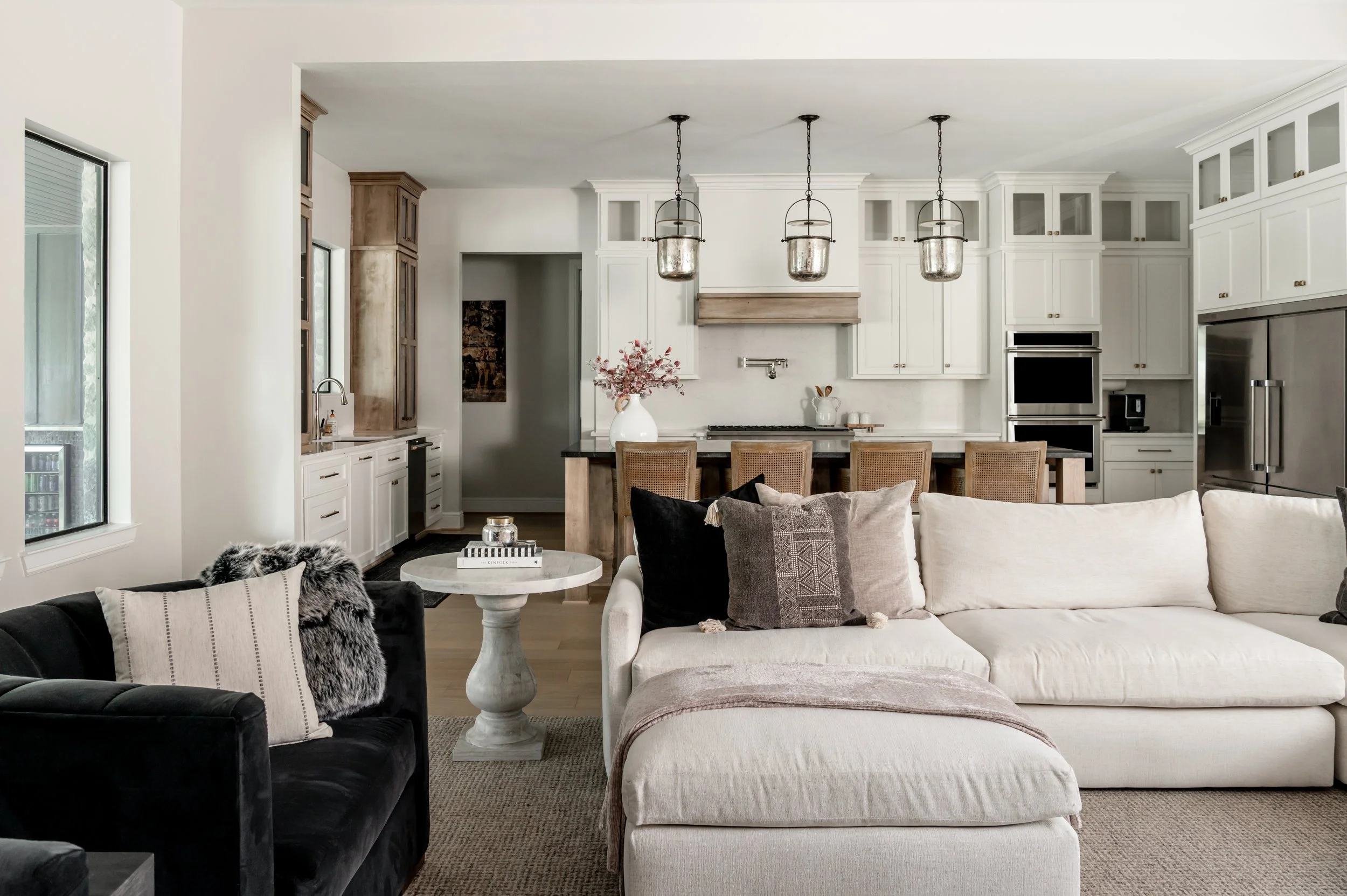 Open-concept living space with white sectional sofa, black armchair, coffee table, and kitchen area with island, white cabinets, and hanging pendant lights.