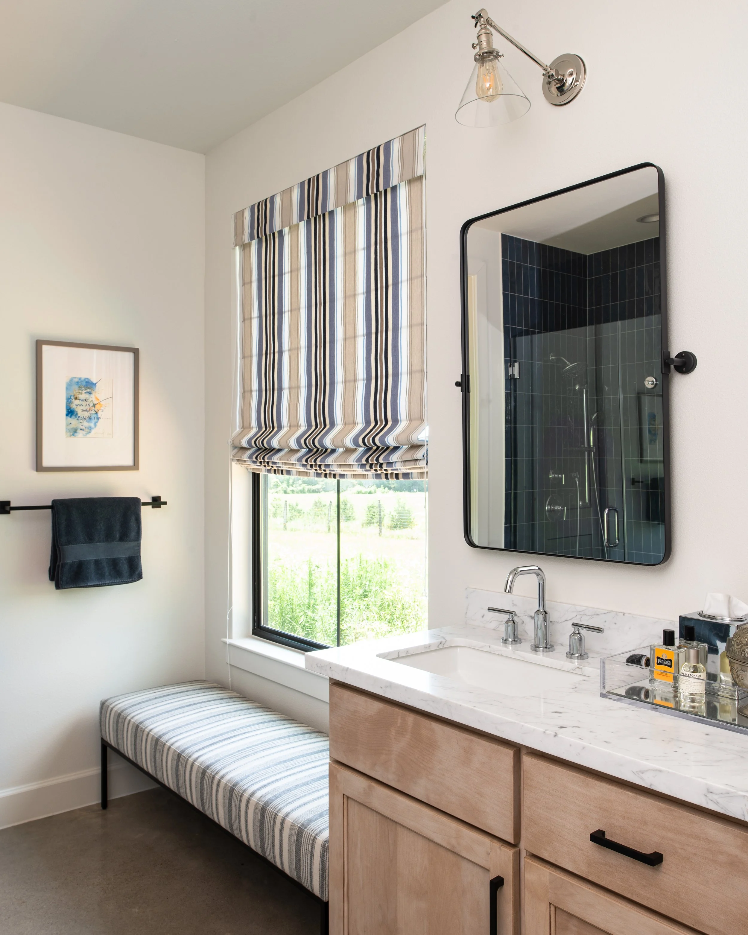Bathroom with striped window treatment, marble countertop, wooden cabinet, black framed mirror, and a wall light fixture.