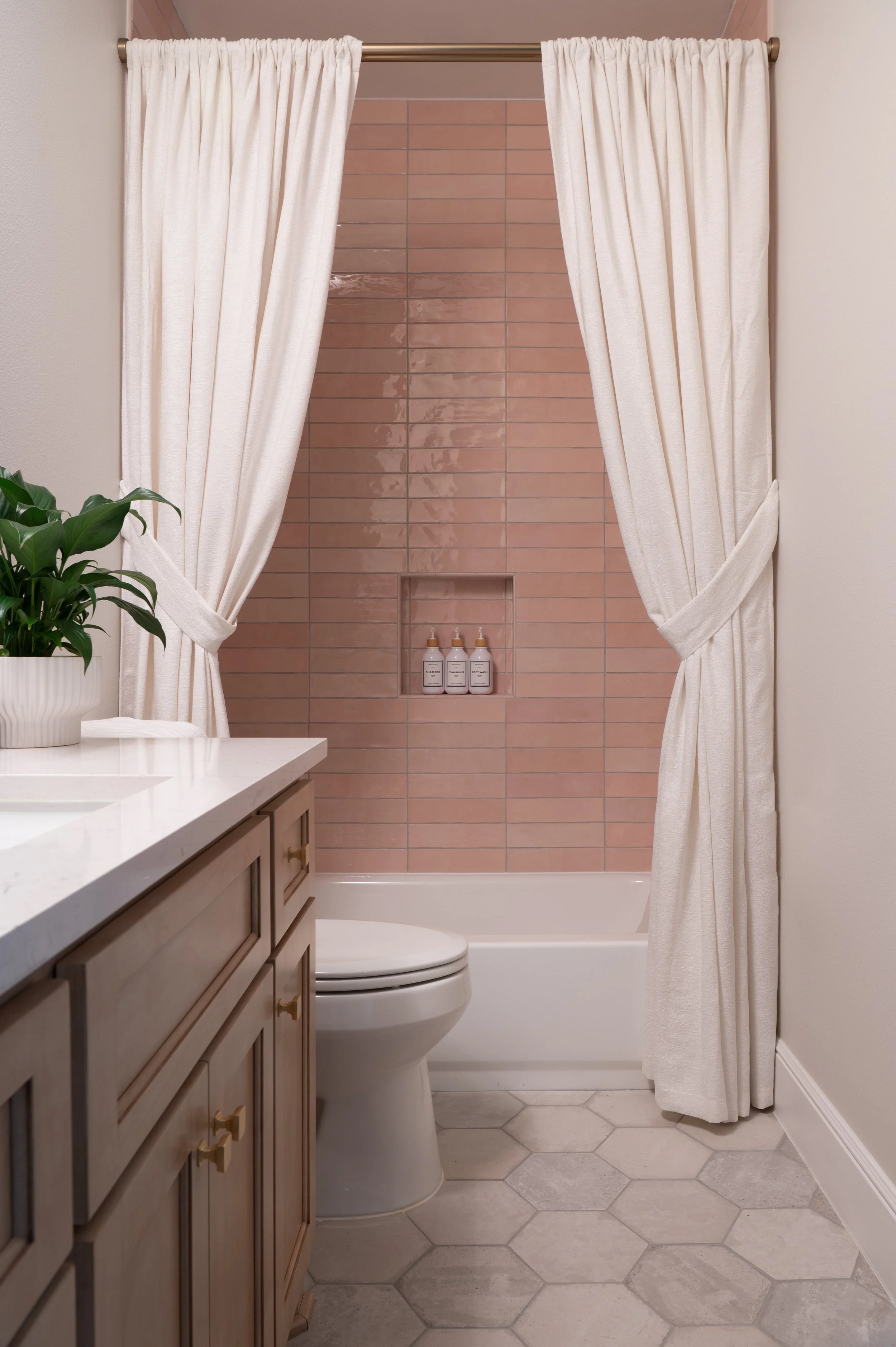 A bathroom with a bathtub enclosed by cream curtains, a pink tiled wall inside the shower, a built-in shelf with three bottles, a toilet, a white countertop with a potted plant, and hexagonal gray floor tiles.