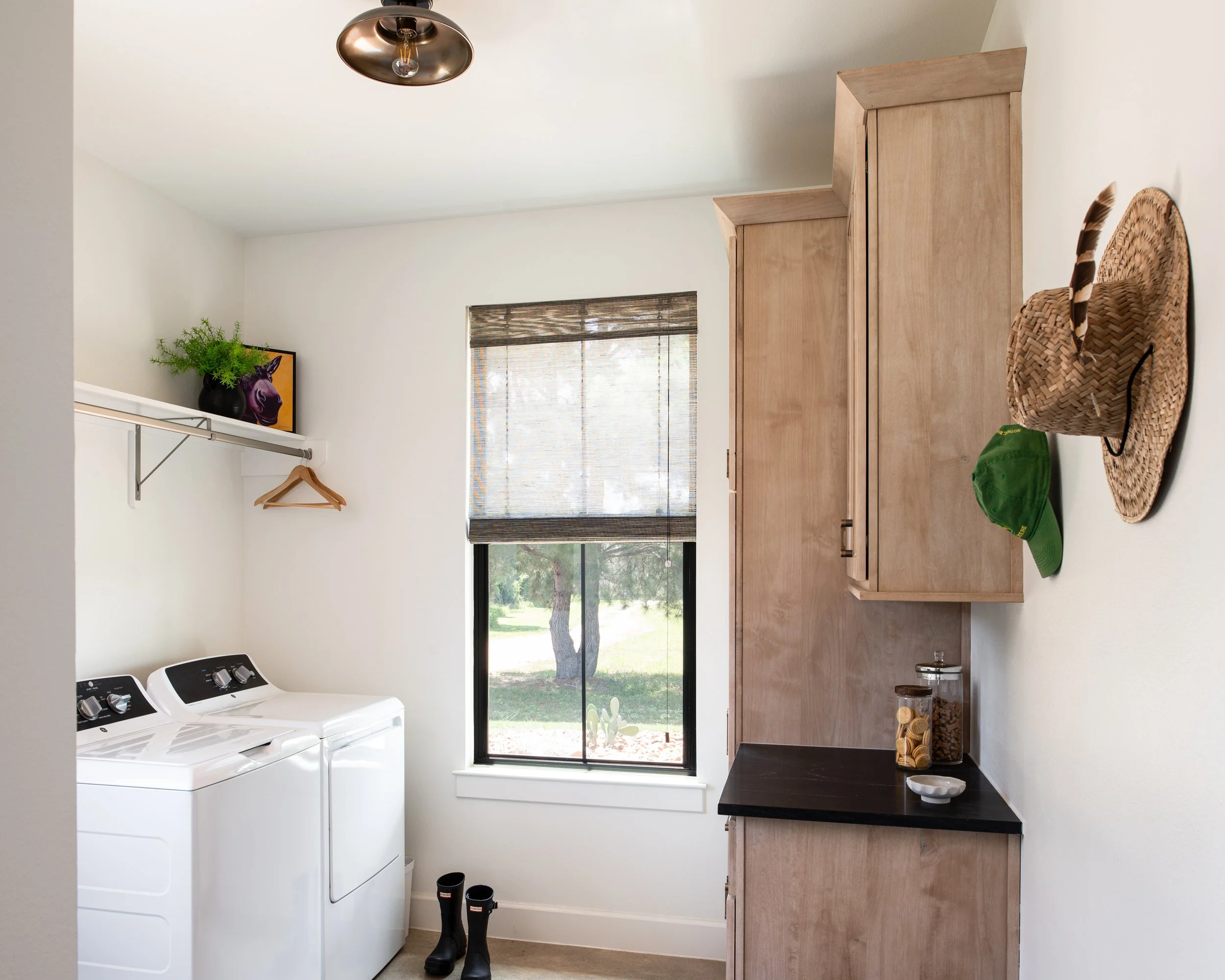 A laundry room with a washing machine, a window with a bamboo shade, wooden cabinets, a shelf with a potted plant and a picture, a straw hat, a green cap, a jar of cookies, and rain boots.