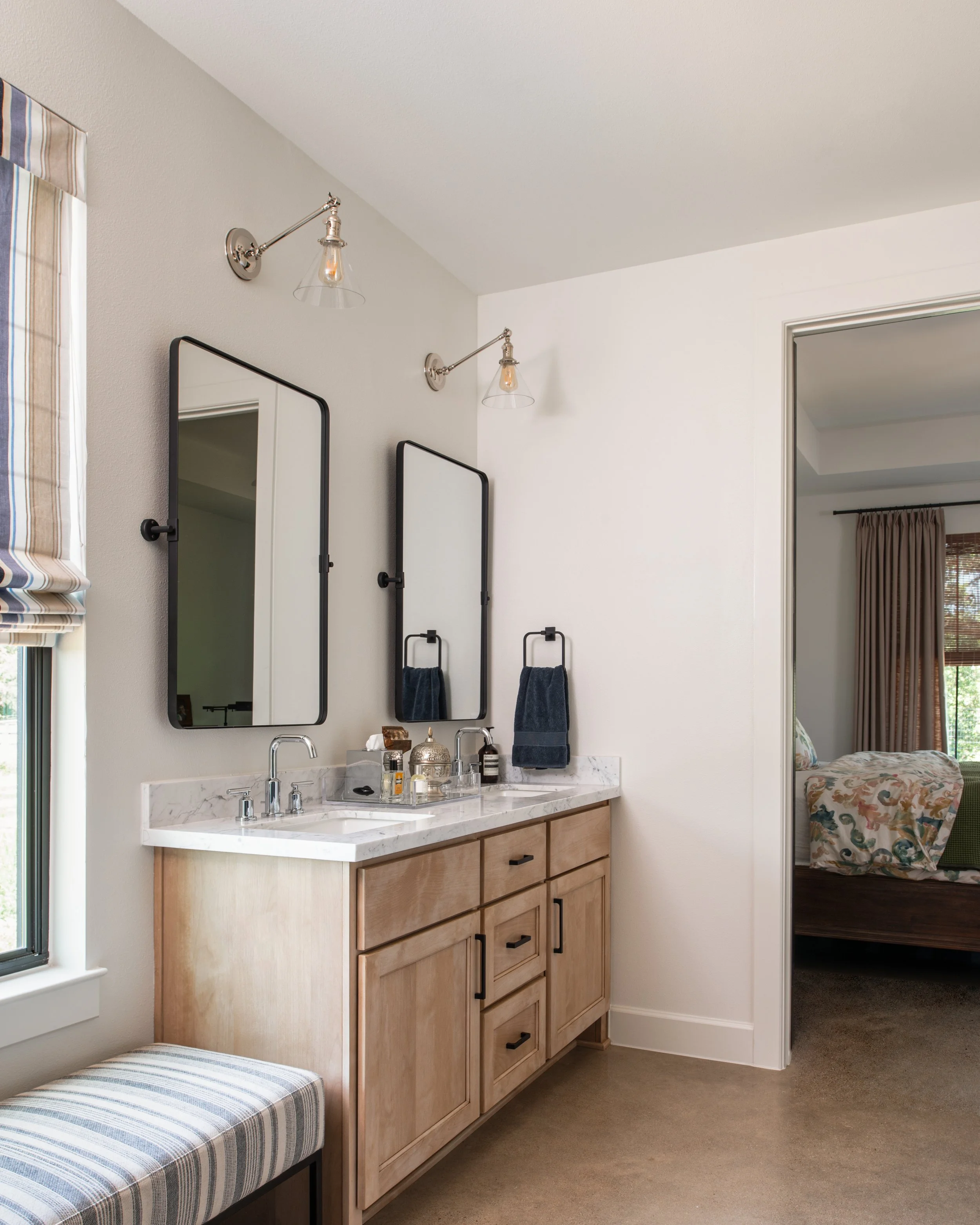 Bathroom vanity with two mirrors, marble countertop, two sinks, and black fixtures. There are navy towels, a window with striped blinds, and a doorway leading to a bedroom with patterned bedding.
