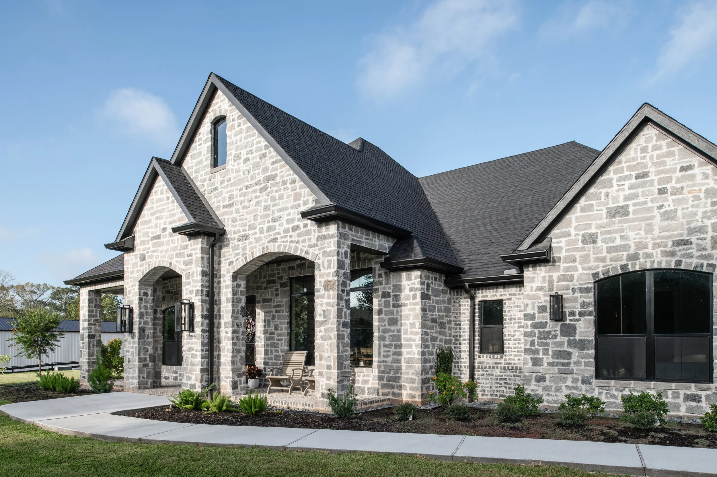 Stone house with black roof, front porch with chair, landscaped yard, and a blue sky with some clouds.