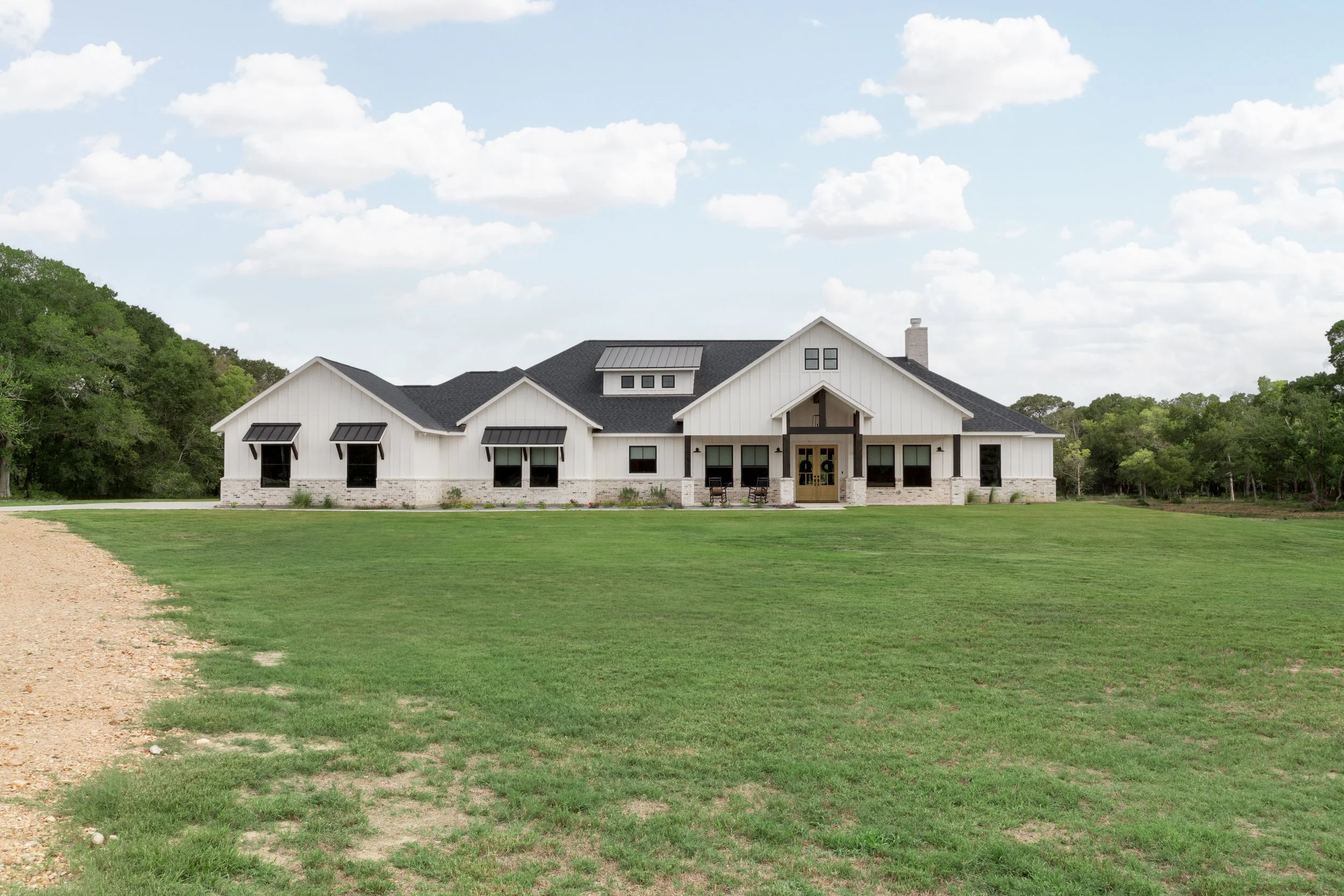 Large white modern farmhouse with black roof, multiple windows including three with black awnings, front porch with two chairs, set on a green lawn with a gravel driveway on the left, surrounded by trees under a partly cloudy sky.