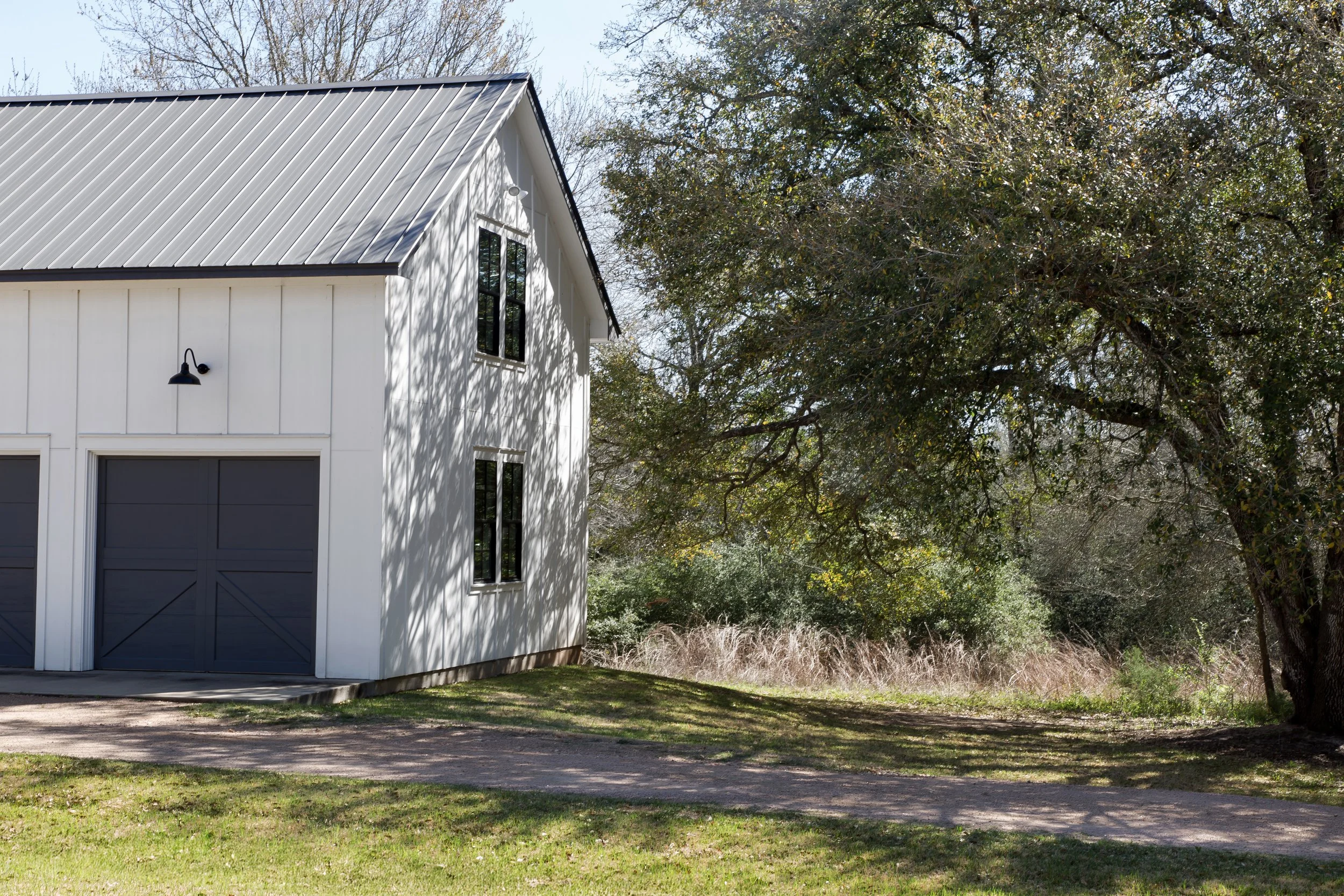 A white modern barn with black doors and windows, black outdoor wall light, surrounded by greenery and tall trees, with shadows on the ground.