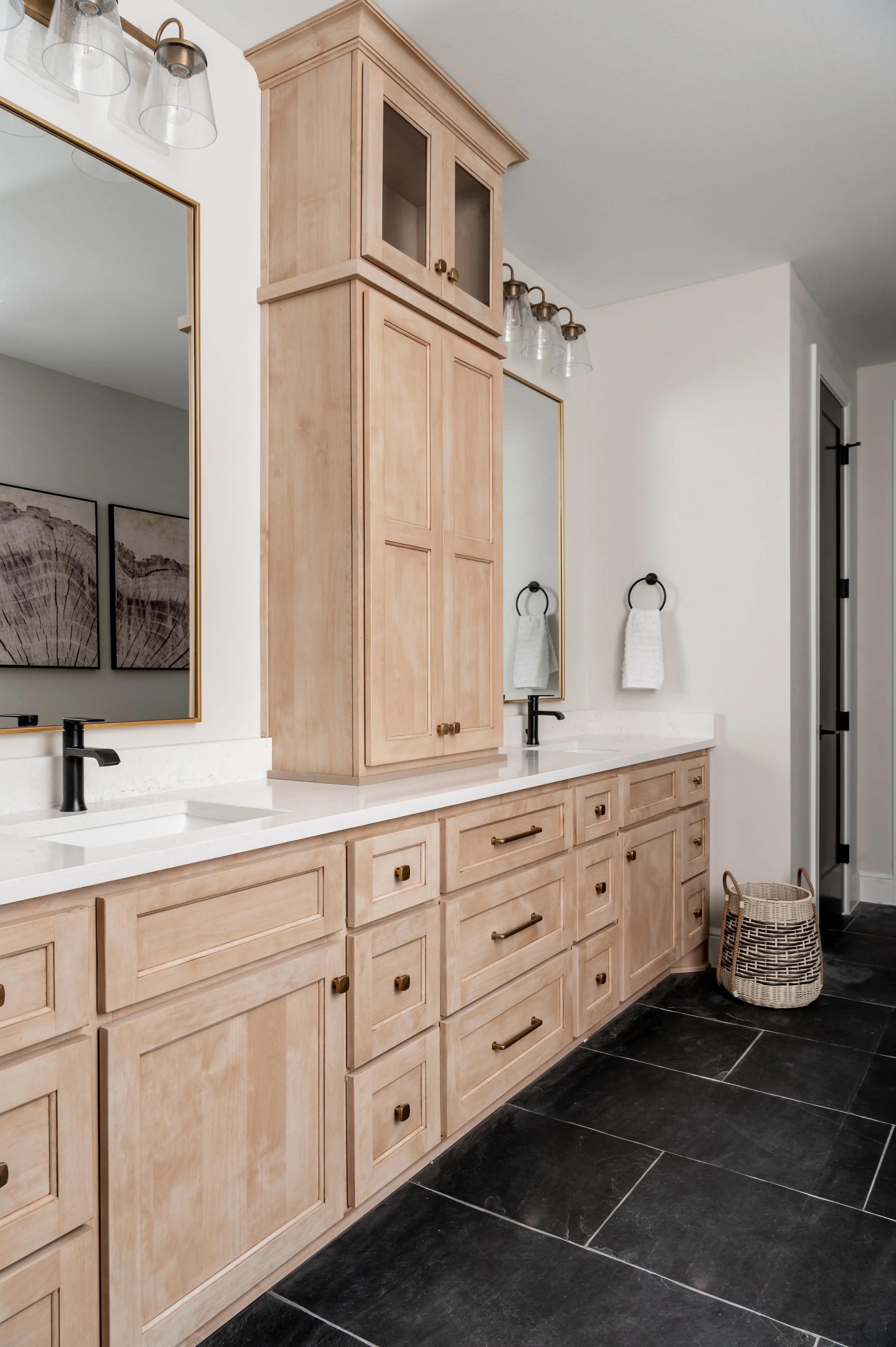 Modern bathroom with light wood cabinetry, white countertop, black fixtures, large mirror, baskets, and dark tiled floor.
