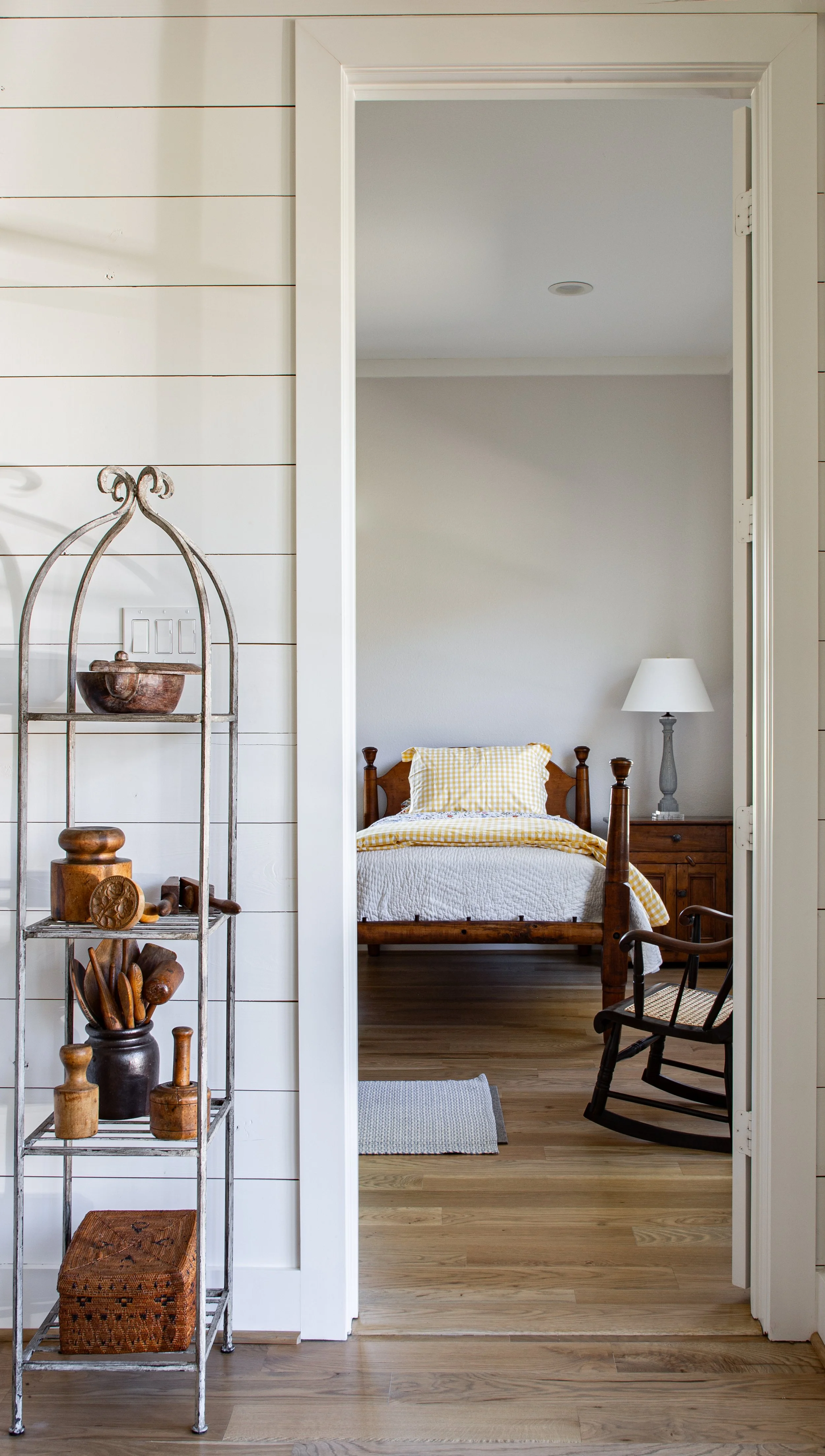 A view through an open doorway into a bedroom with a wooden bed, yellow checkered bedding, a nightstand with a lamp, a rocking chair, and a small gray rug on wooden flooring, with a metal shelf holding wooden kitchen utensils on the left.