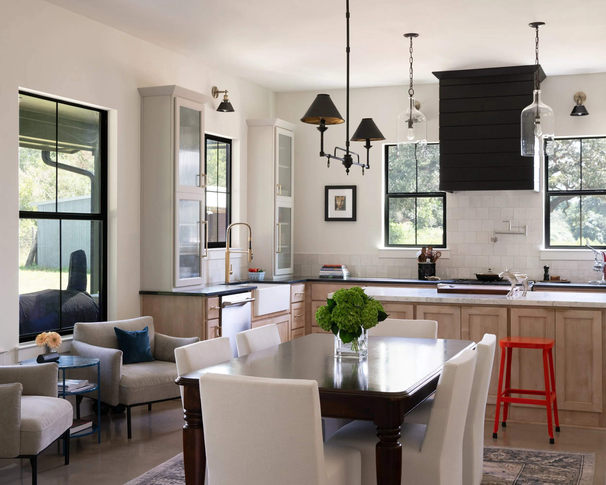 Open-concept kitchen and dining area with white cabinets, black countertops, large windows, and modern lighting fixtures. There's a wooden dining table with white chairs, a sofa with pillows, and a red stool next to the kitchen counter.