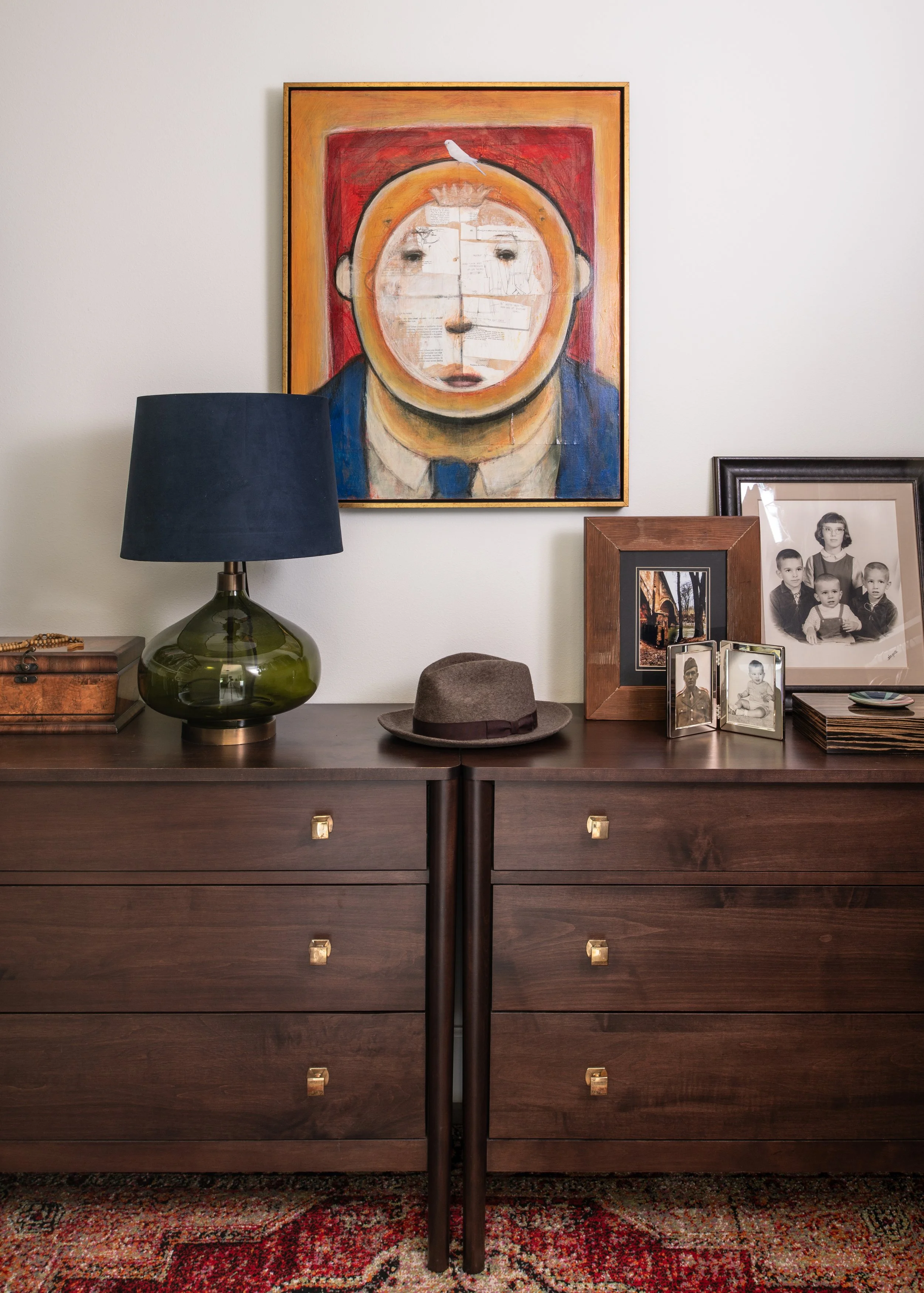 A dark wood dresser with brass square knobs, topped with a brown fedora hat, a green glass lamp with a black shade, framed family photos, a wooden picture frame with a street scene, and a stack of black coasters. Behind the dresser on the wall is a v