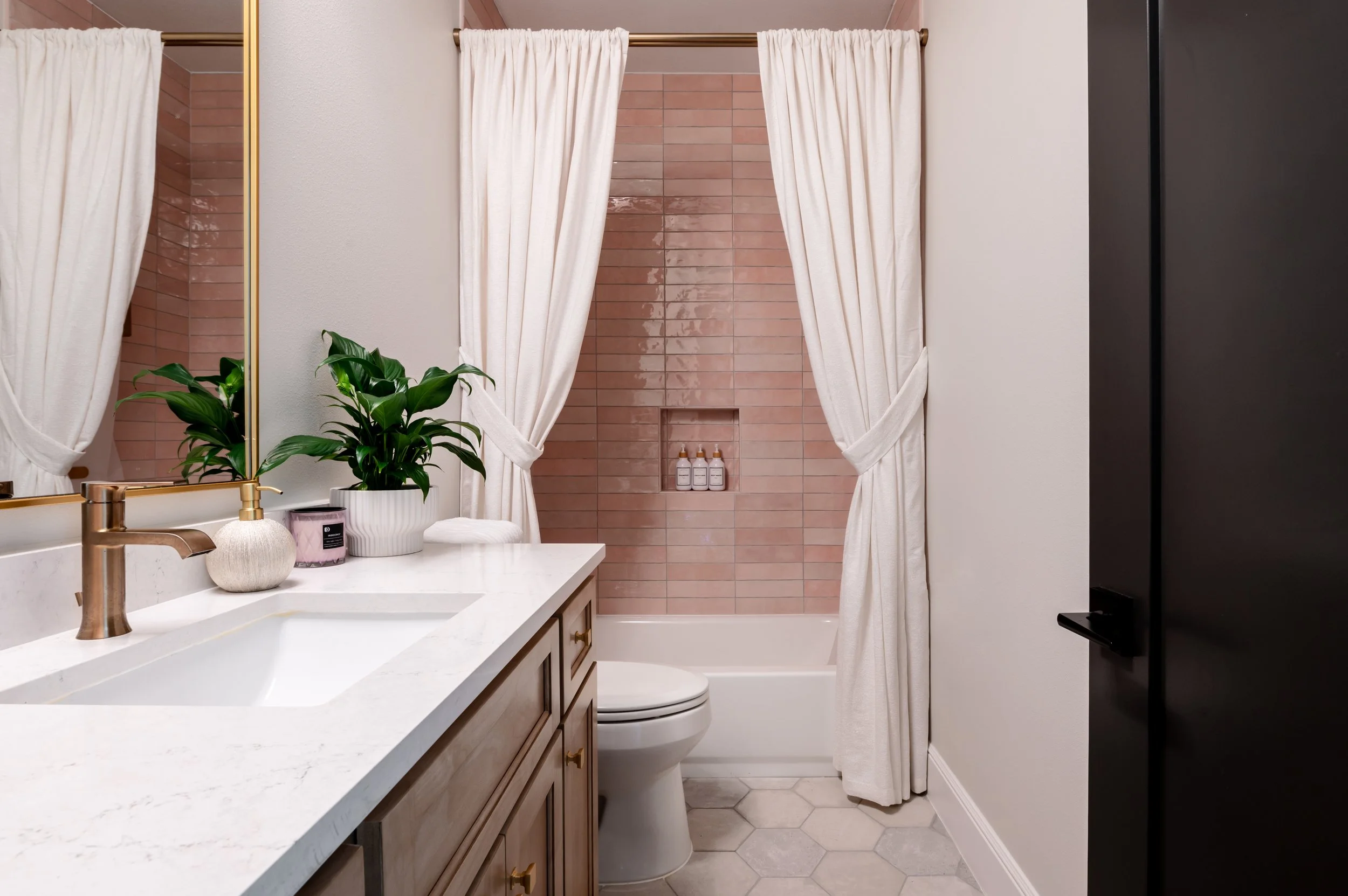 Bathroom with a white vanity, a green plant, a mirror, a bathtub with pink tiles and white shower curtain, toilet, and black cabinet.