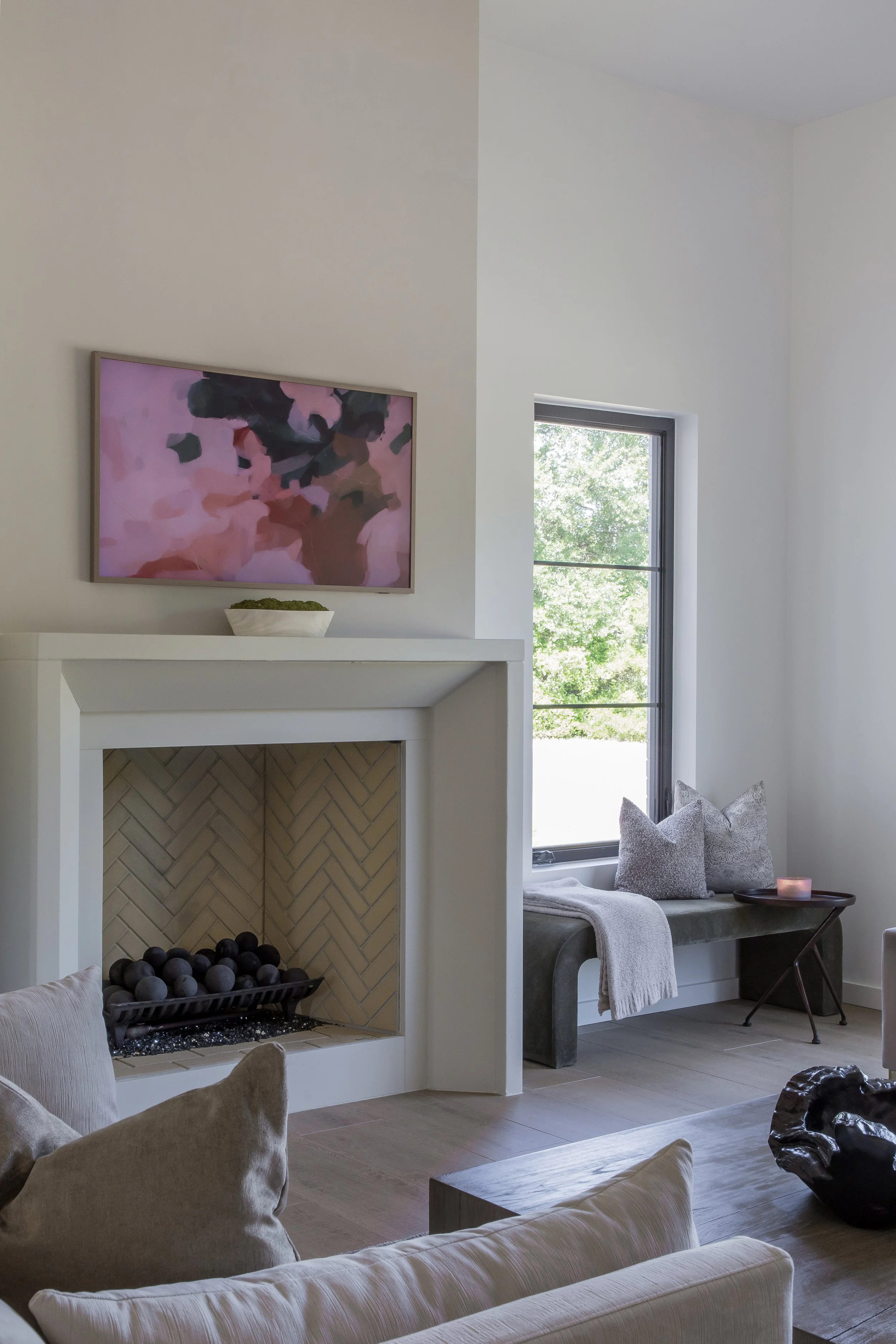 A cozy living room with a white fireplace, abstract pink and black artwork above it, natural light coming through a window with greenery outside, beige sofa with cushions, and a small table with a candle.