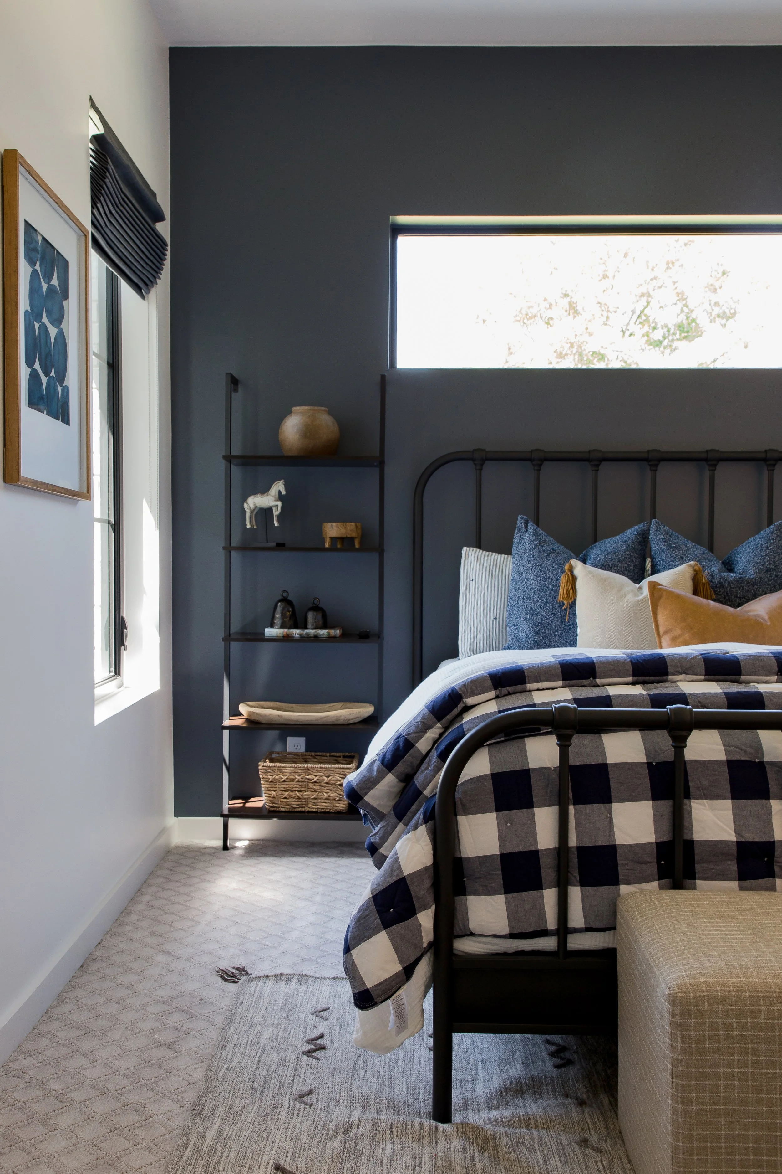 A bedroom with a black metal bed frame, checkered bedding, and decorative pillows, featuring a dark blue accent wall and a window above the bed that lets in natural light, with a black shelving unit next to the bed holding decorative items.