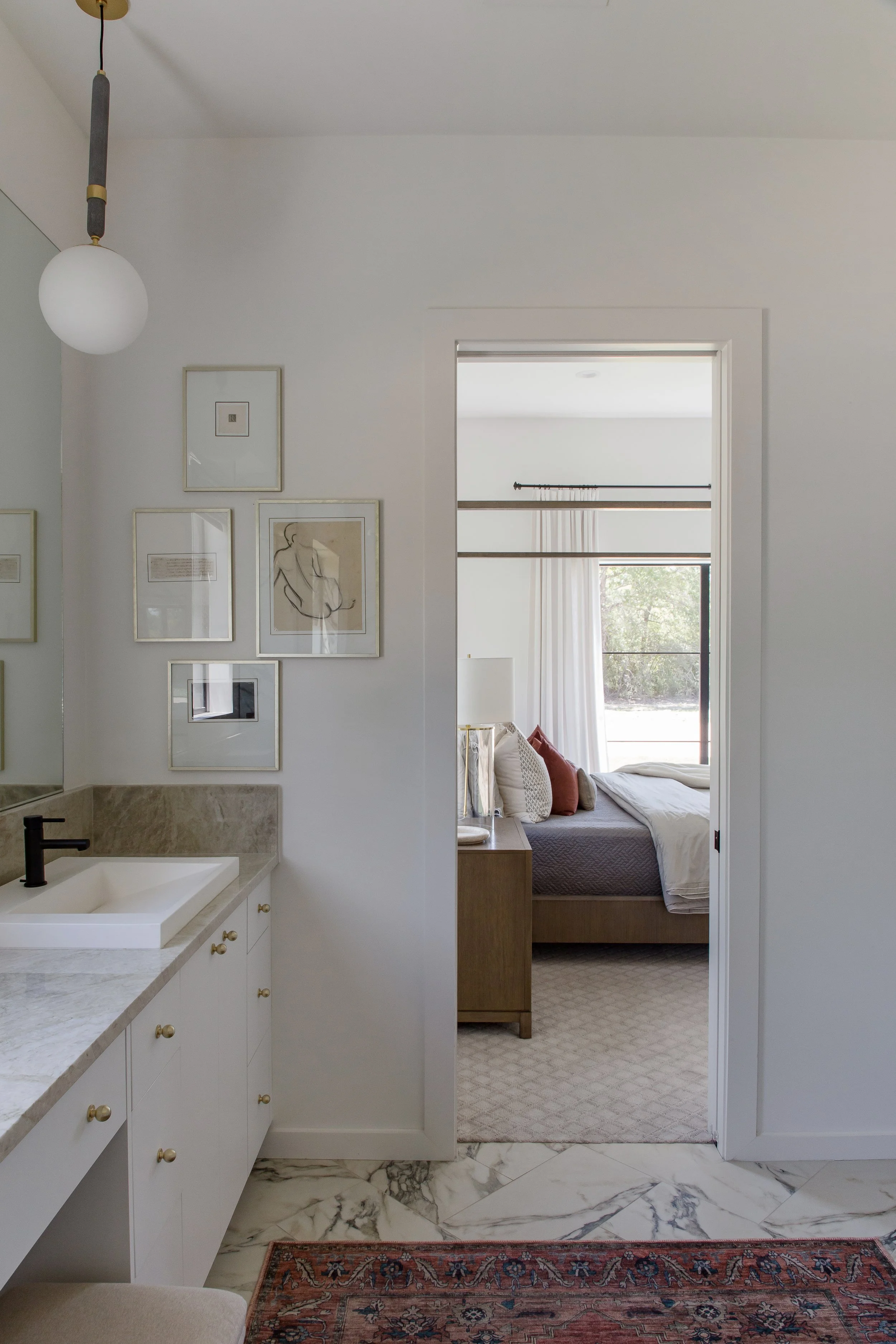 Interior view of a bedroom visible through an open door from a bathroom with marble flooring, a white vanity with a marble countertop, framed artworks on the wall, and a modern hanging light fixture.