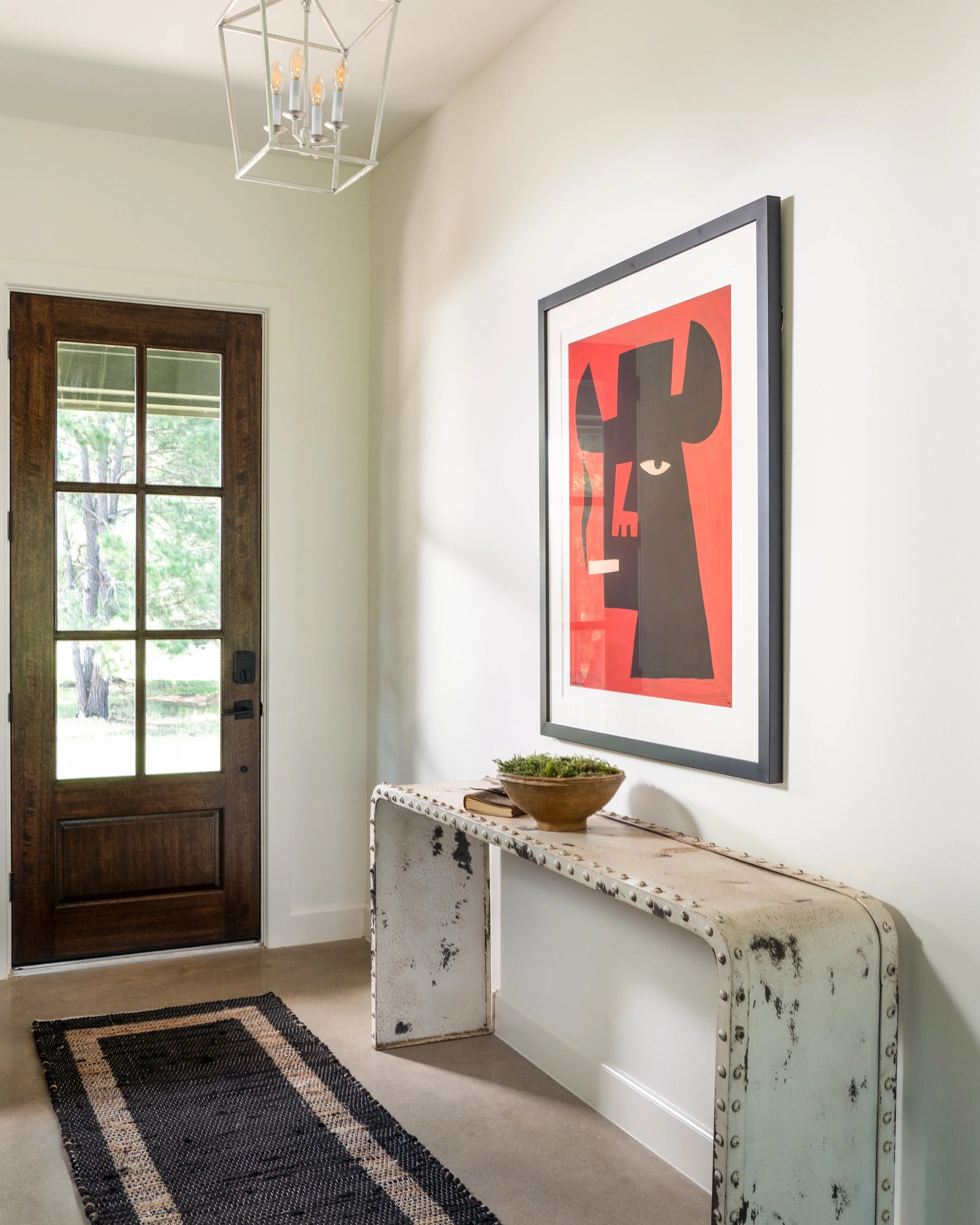 Entrance hallway with a wooden door, a modern metal chandelier, a framed abstract art piece with black and red shapes, a distressed white console table, a bowl with green moss, books, and a black patterned rug.