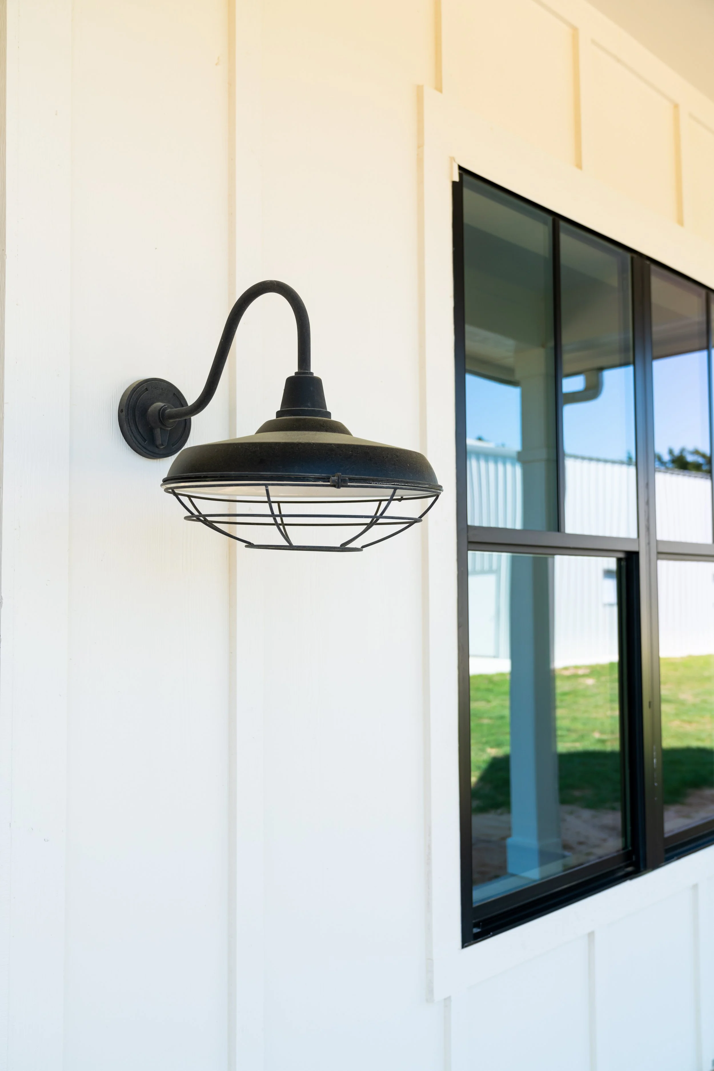 Black outdoor wall light fixture mounted on white wall, near a large window with a view of a grassy yard.