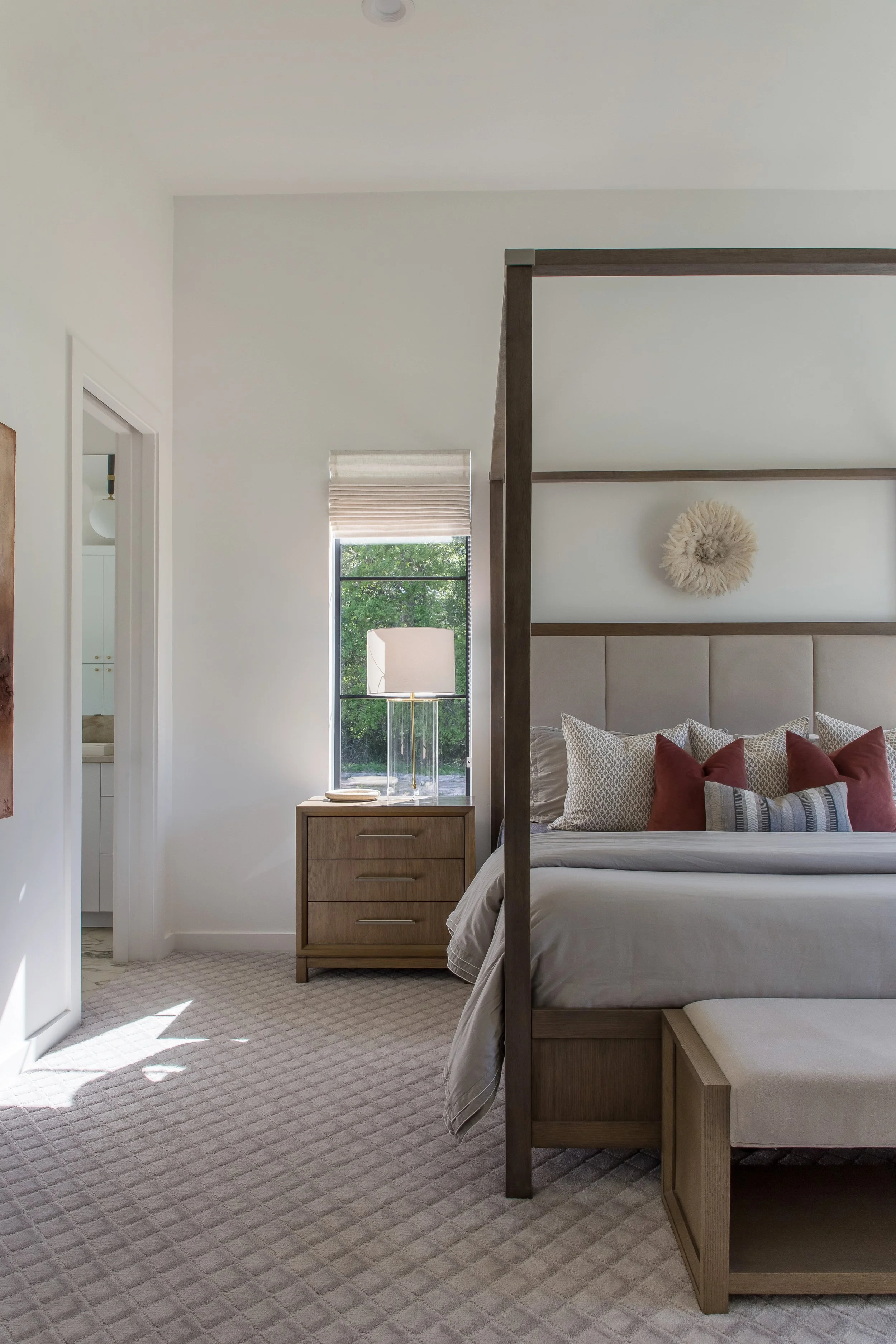 A bedroom with a bed, pillows, a nightstand with a lamp, a window with a view of trees, and a door leading to a bathroom.