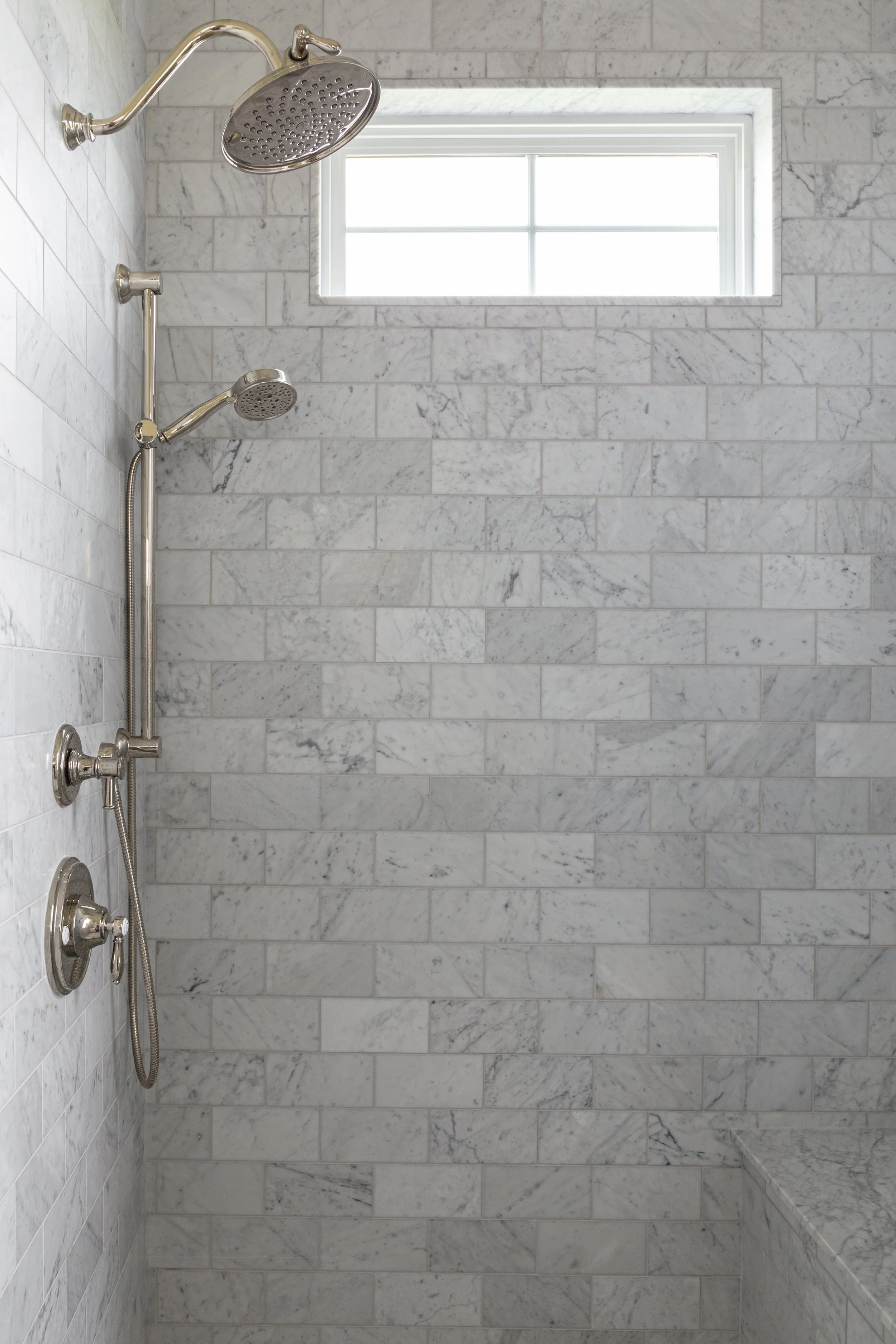 Shower area with marble tile walls, a window, a showerhead, and three control knobs.
