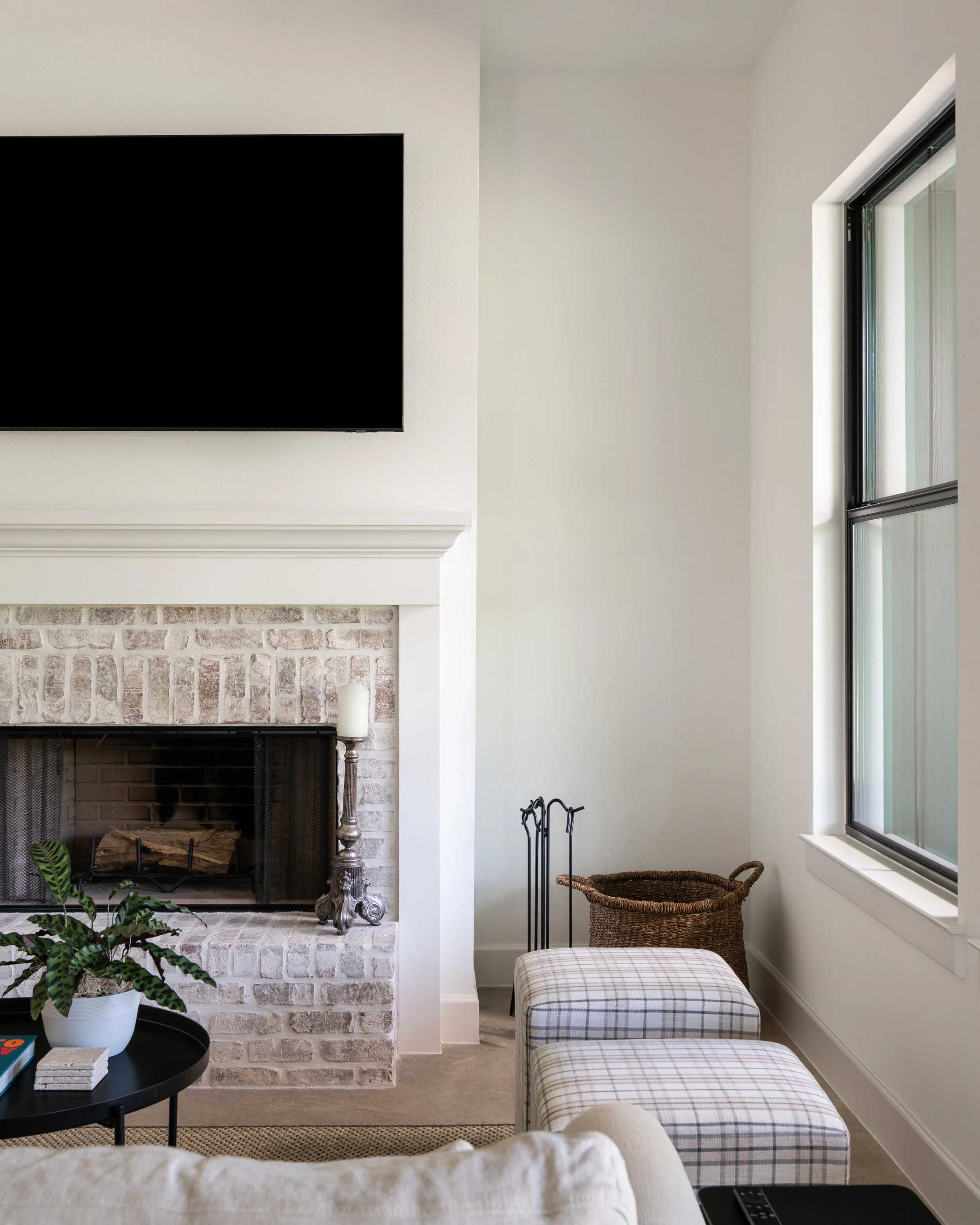 Living room interior with fireplace, large window, plaid ottomans, potted plant, and wall-mounted TV.
