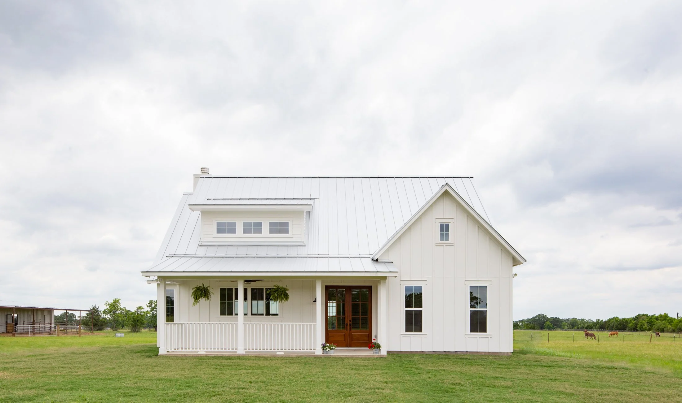 White farmhouse with a metal roof, front porch with hanging plants, wooden door, and surrounded by green grass and open fields with horses.