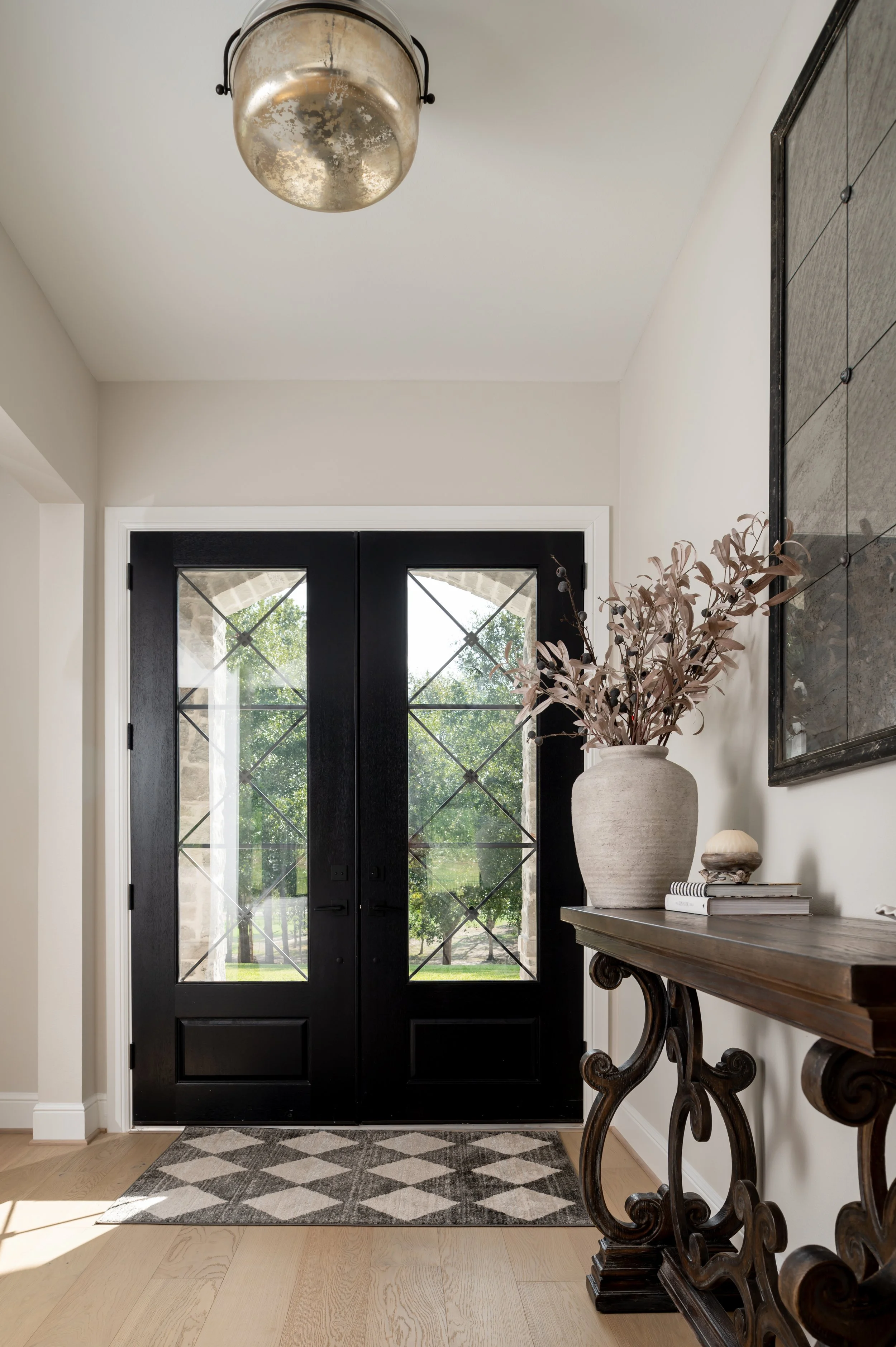 Entryway with black double doors, glass panes, view of greenery outside, decorative console table with vase filled with dried branches, and a framed artwork.