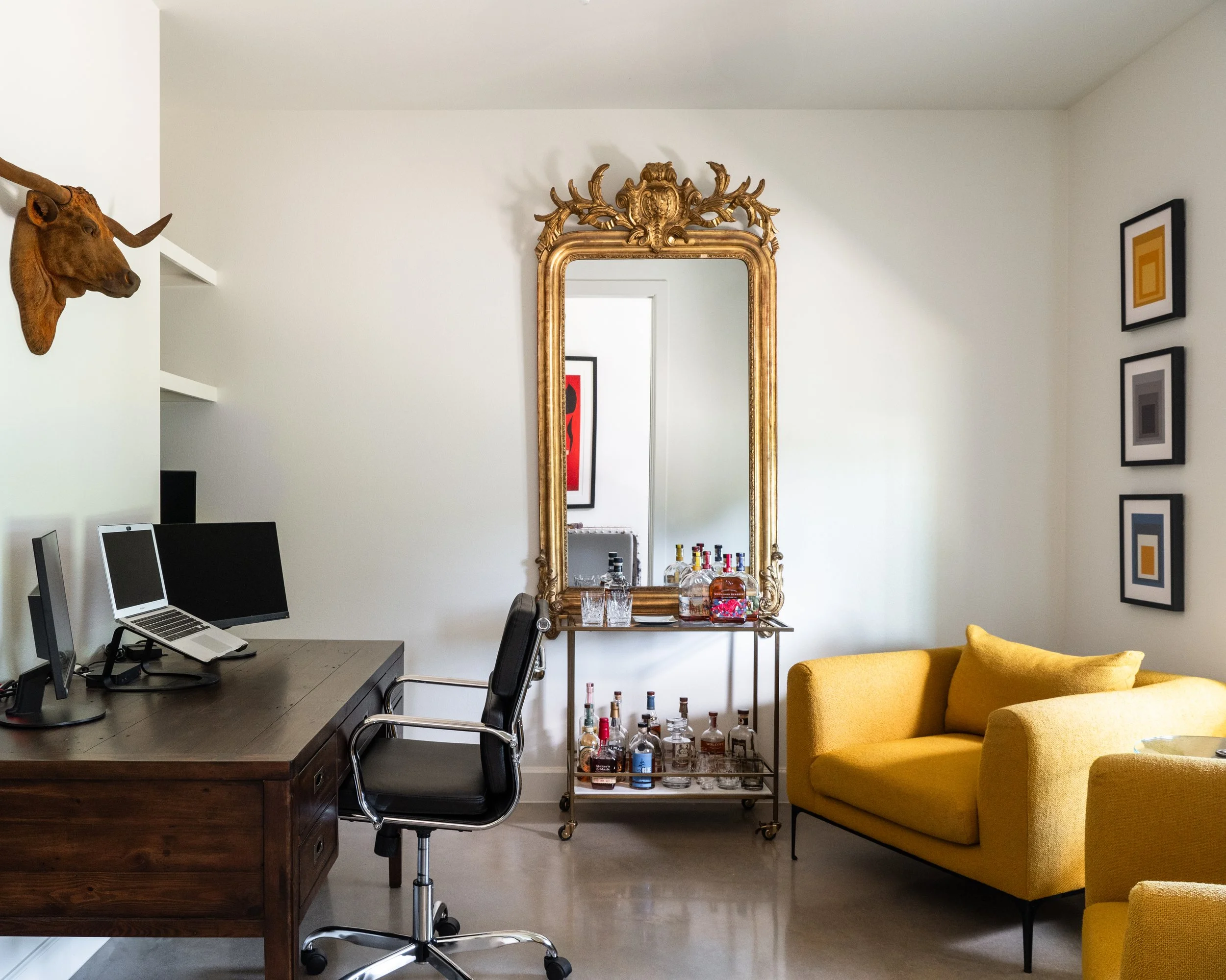 Living room with white walls, gold ornate mirror, yellow sofa, black office chair, wooden desk with dual monitors, bar cart with various liquor bottles, and framed artwork on the wall. A mounted bull head decorates the corner.