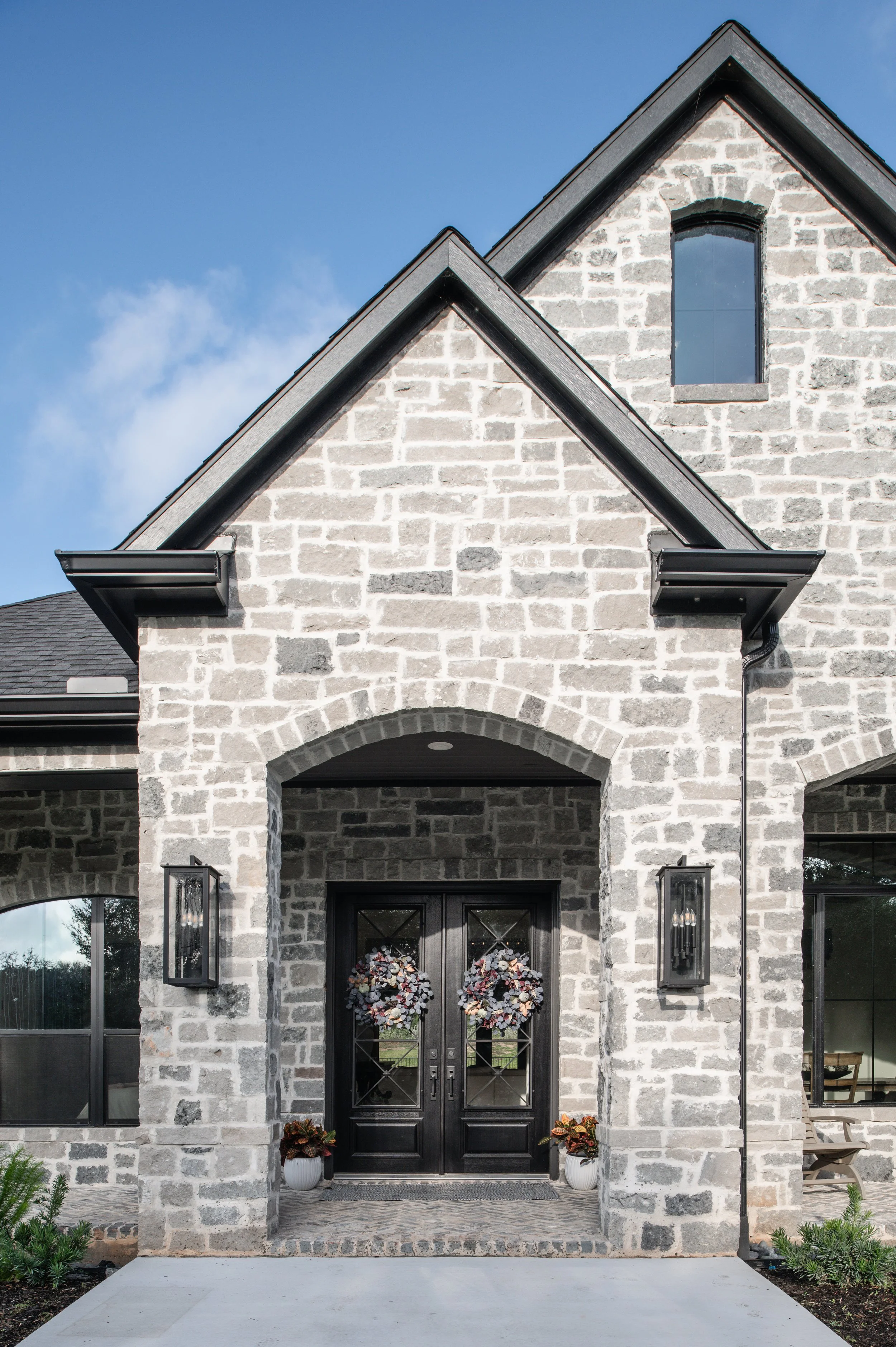 Modern stone house with black door and wreaths, large windows, and outdoor lighting.
