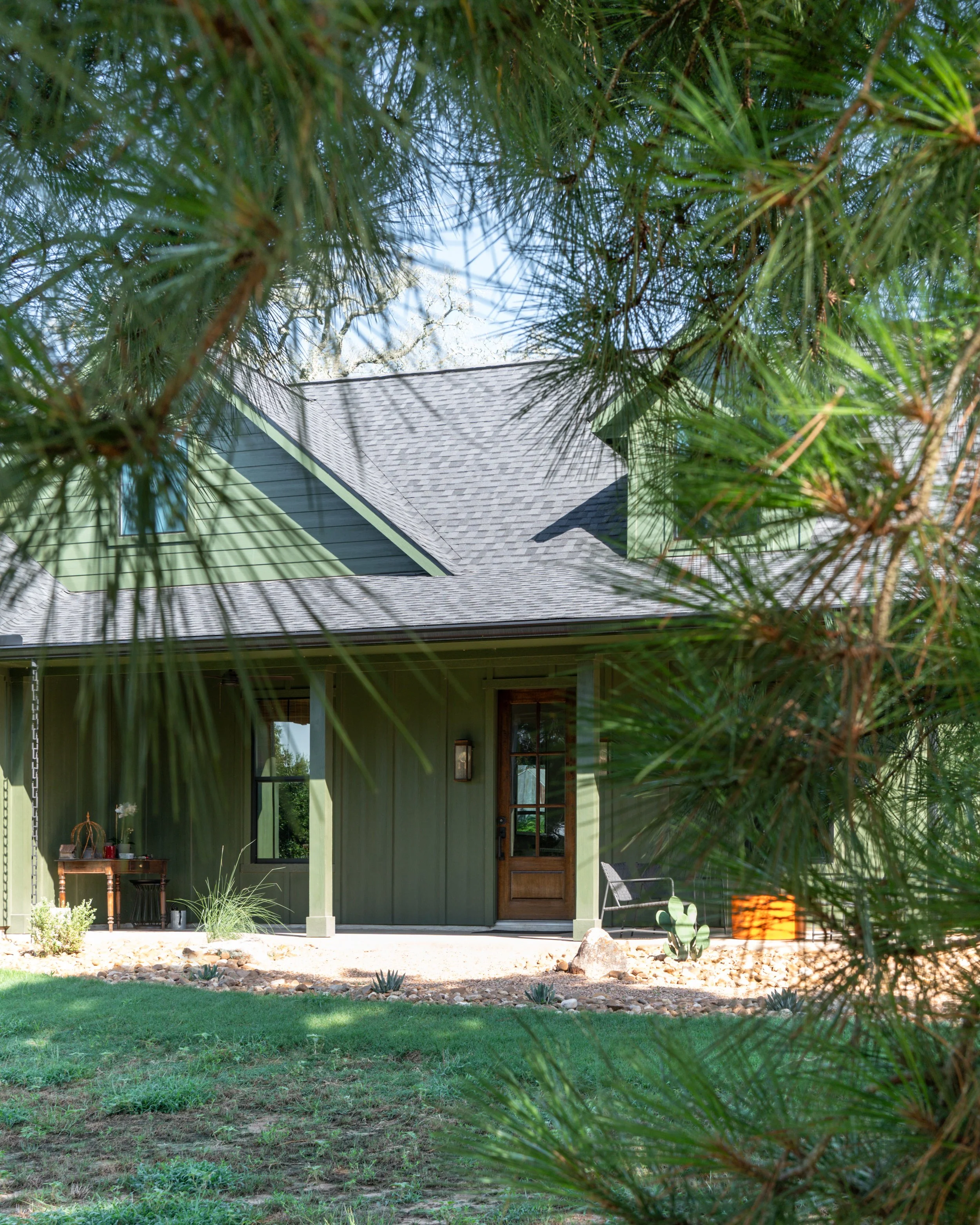 A house painted green with a front porch and wooden door, framed by pine tree branches in the foreground, with a lawn and desert plants in front.