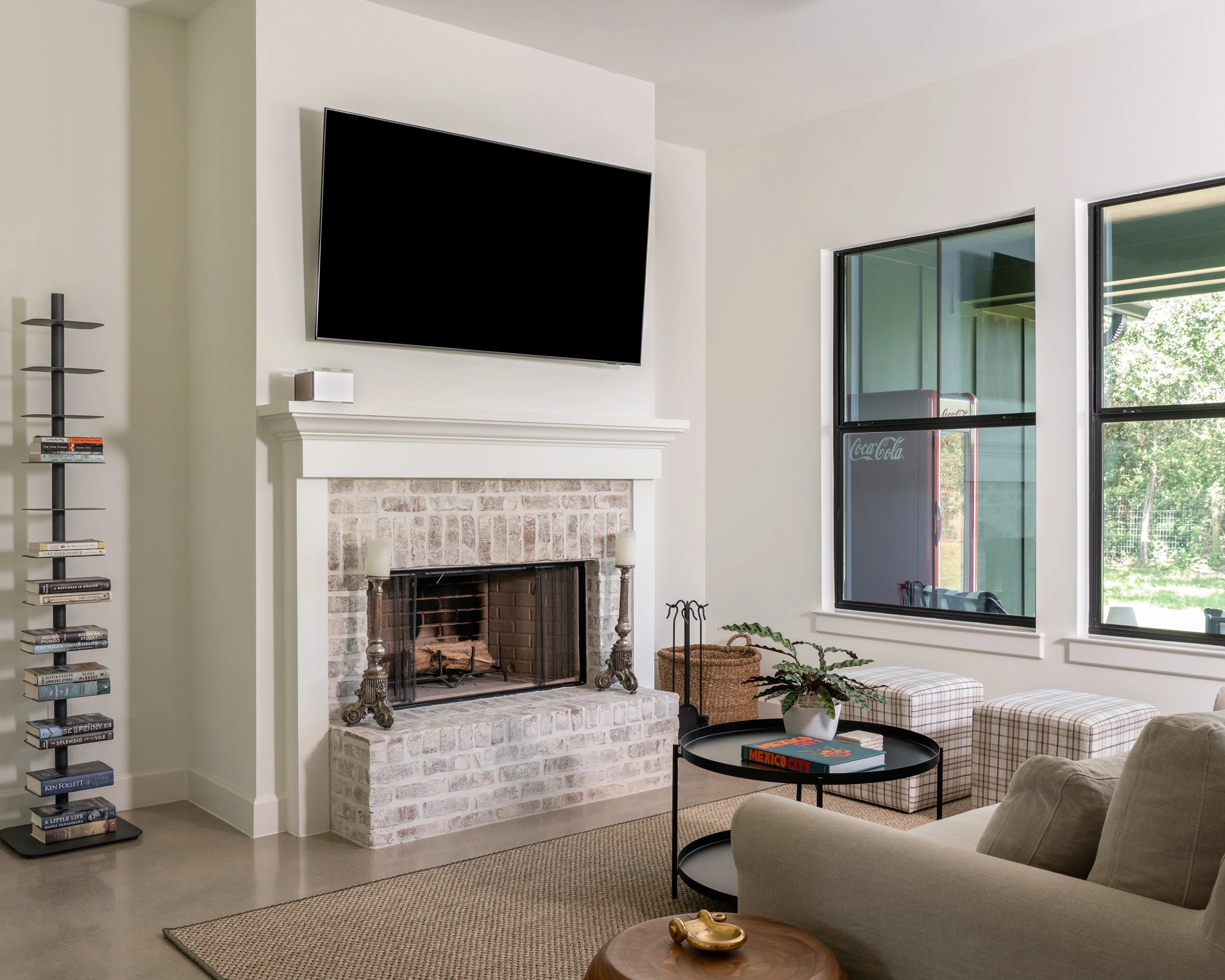 Living room with brick fireplace, wall-mounted TV, beige sofa, plaid ottomans, black round coffee table with book, window showing Coca-Cola vending machine outside, potted plant, and tall black bookshelf.