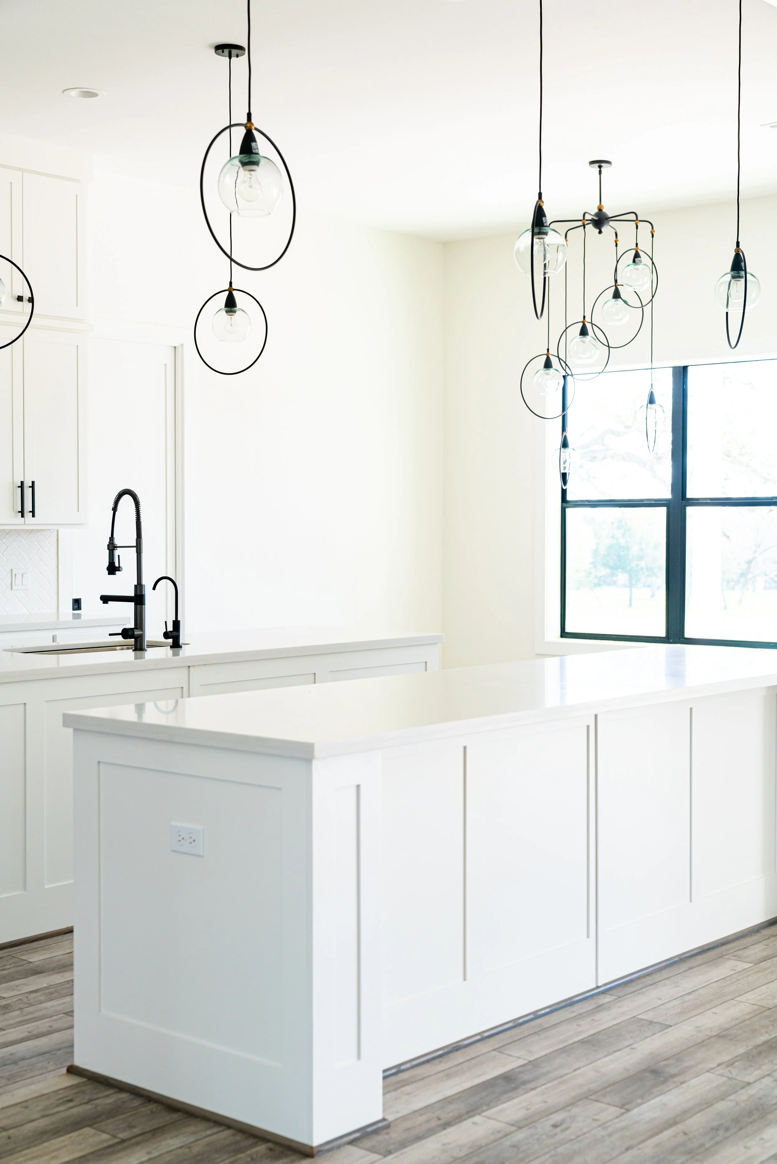 Modern kitchen with white cabinets, a kitchen island, black faucets, large window, and contemporary pendant lights.