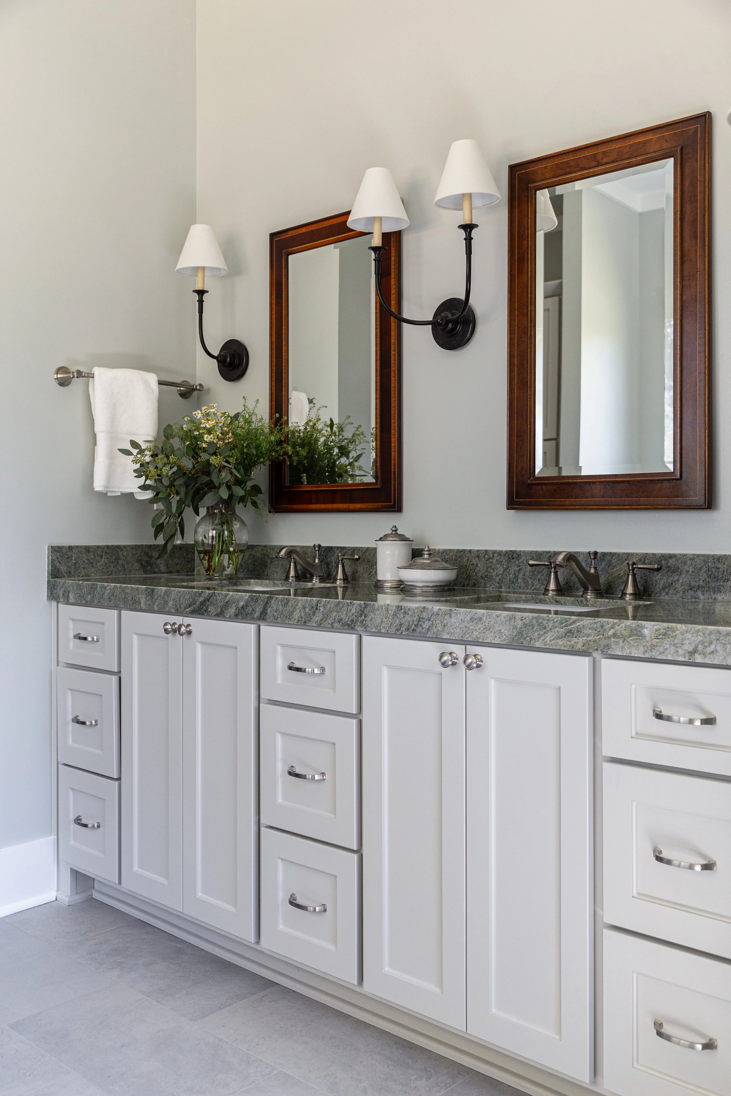 A bathroom vanity with white cabinets, a grey countertop, two mirrors, wall-mounted lights, a towel rack, and a vase with flowers.