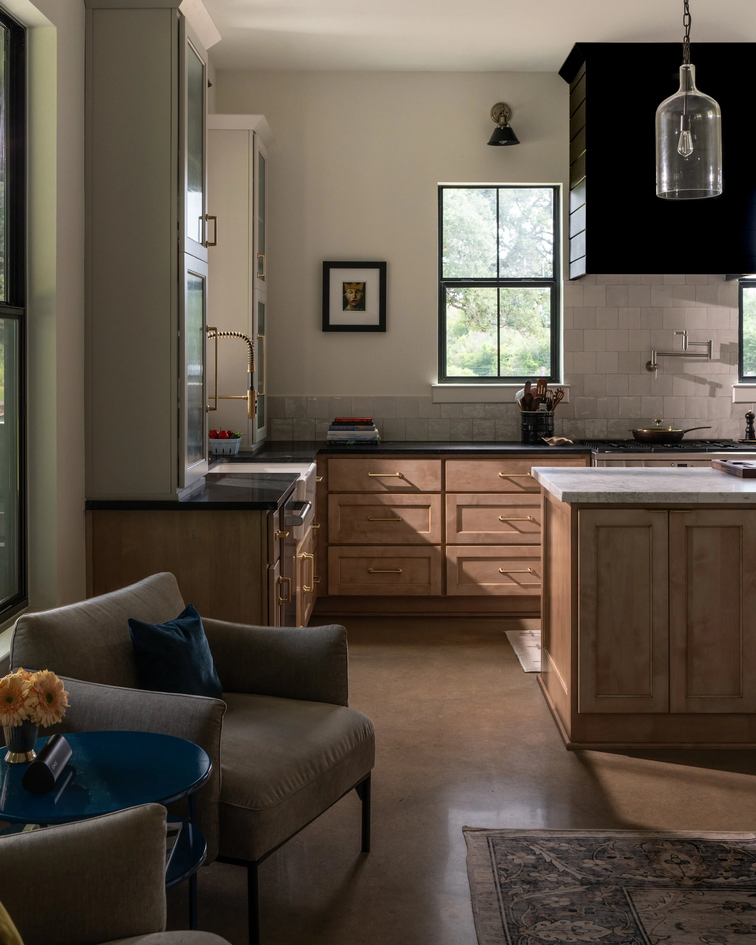 A modern kitchen with wooden cabinets, black countertops, and a white tiled backsplash. There are two windows, one in the center and one near the stove. A black pendant light hangs from the ceiling. A small sofa with a side table and flowers is in th
